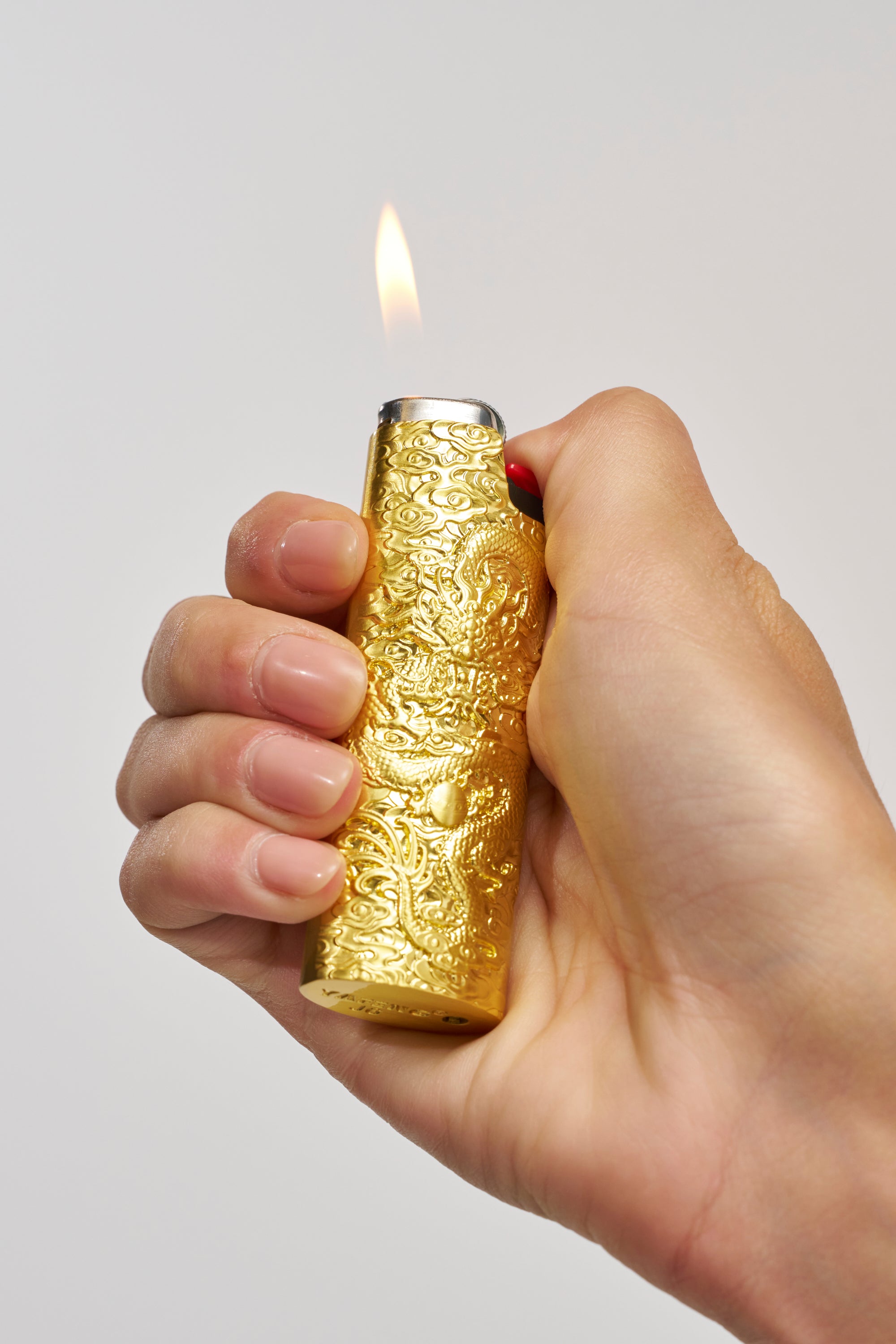 A hand holds the GOLDEN ERA LIGHTER CASE, a gold-tone accessory with engraved detailing, as it produces a small flame against a simple light background.
