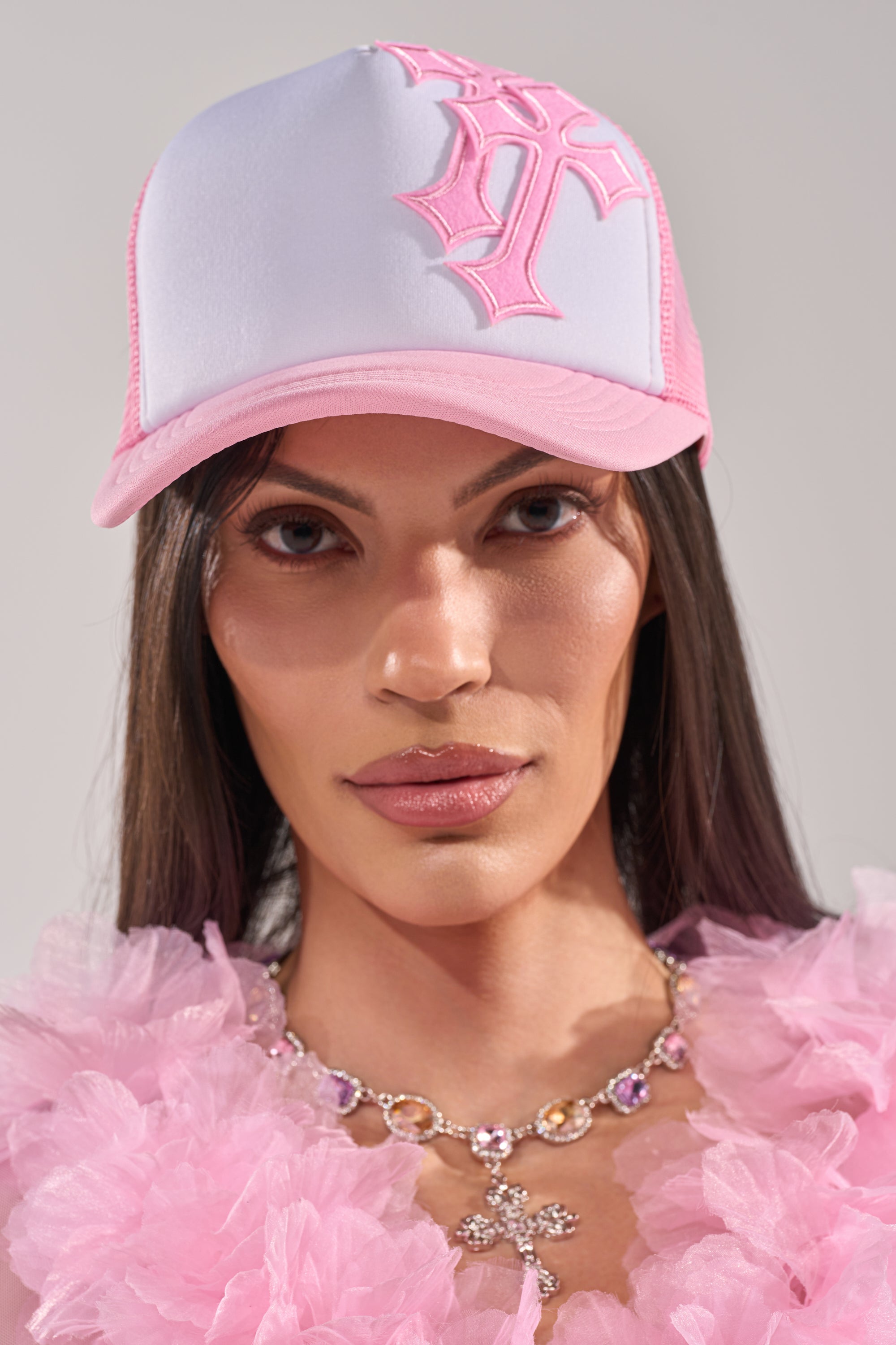 A person with long straight hair wears the CROSS ME PATCH HAT, a pink ruffled top, and a jeweled cross necklace against a neutral background. 2