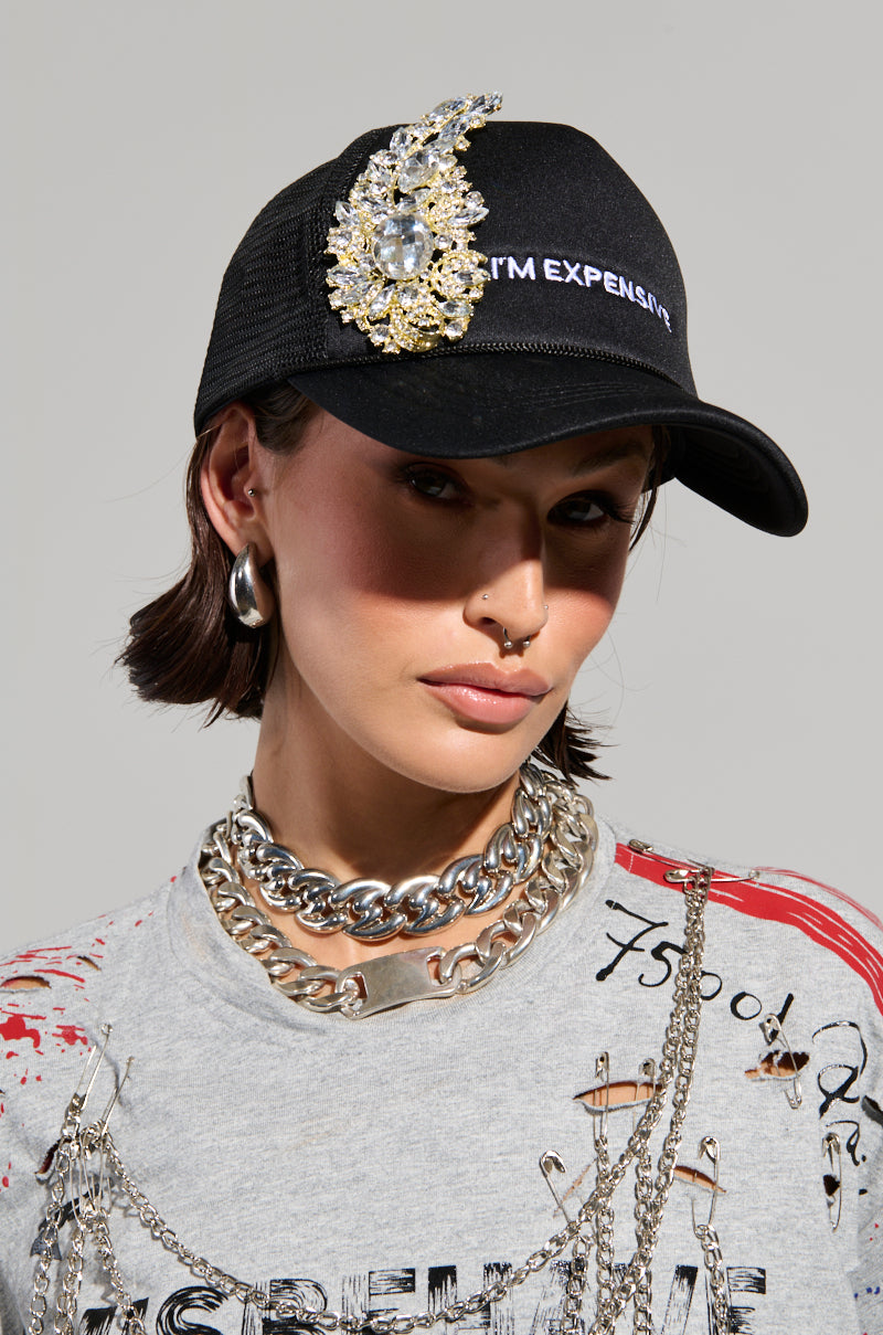 A person wearing the stylish EXPENSIVE TASTE TRUCKER HAT confidently displays their expensive taste. They pair it with short hair, silver earrings, a layered chain necklace, and a graphic t-shirt featuring bold red accents.