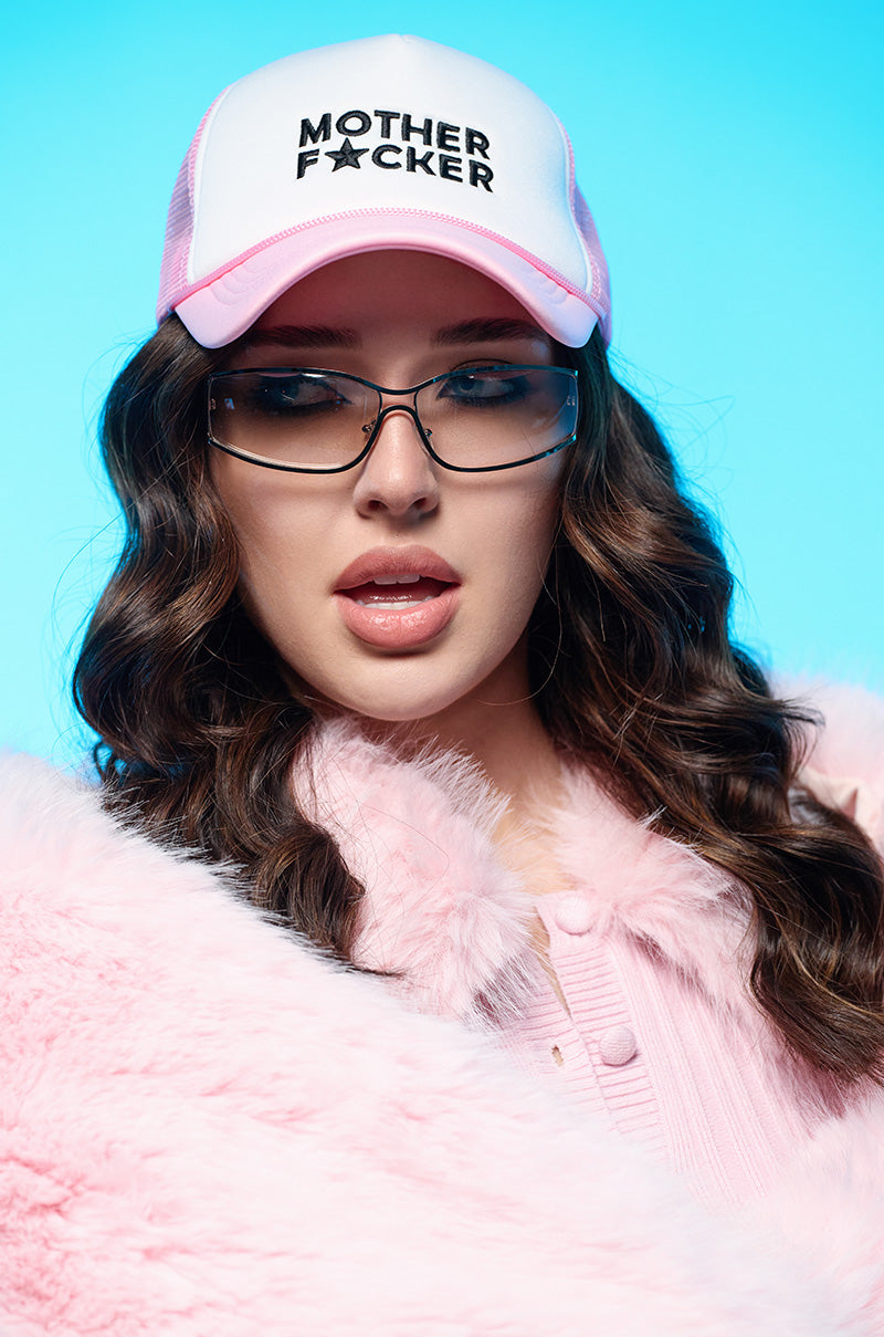 A person with long brown wavy hair is wearing glasses and the MOTHER TRUCKER HAT IN PINK, featuring black embroidered text. They're dressed in a pink fluffy jacket against a bright blue background, with their expression focused forward.