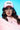A person with long brown wavy hair is wearing glasses and the MOTHER TRUCKER HAT IN PINK, featuring black embroidered text. They're dressed in a pink fluffy jacket against a bright blue background, with their expression focused forward.