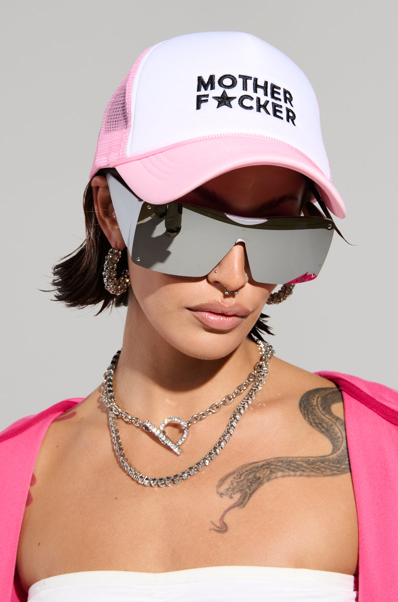 A person sporting the MOTHER TRUCKER HAT IN PINK, complemented by large reflective sunglasses, and accessorized with silver jewelry like a chain necklace and hoop earrings. They display an arm tattoo and wear a pink outfit with an adjustable snapback closure.