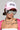 A person sporting the MOTHER TRUCKER HAT IN PINK, complemented by large reflective sunglasses, and accessorized with silver jewelry like a chain necklace and hoop earrings. They display an arm tattoo and wear a pink outfit with an adjustable snapback closure.