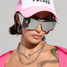A person sporting the MOTHER TRUCKER HAT IN PINK, complemented by large reflective sunglasses, and accessorized with silver jewelry like a chain necklace and hoop earrings. They display an arm tattoo and wear a pink outfit with an adjustable snapback closure.