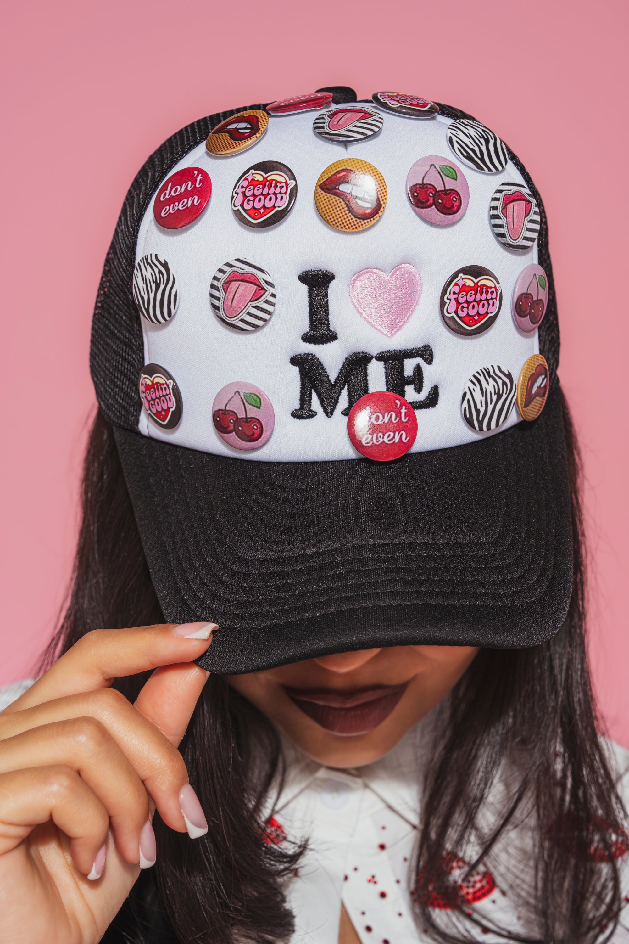 A person wears the LOVE ME BUTTON HAT, a black and white snapback trucker cap adorned with colorful pins and patches like “I ♥ ME,” holding its brim. Only their lower face is visible against a pink background.