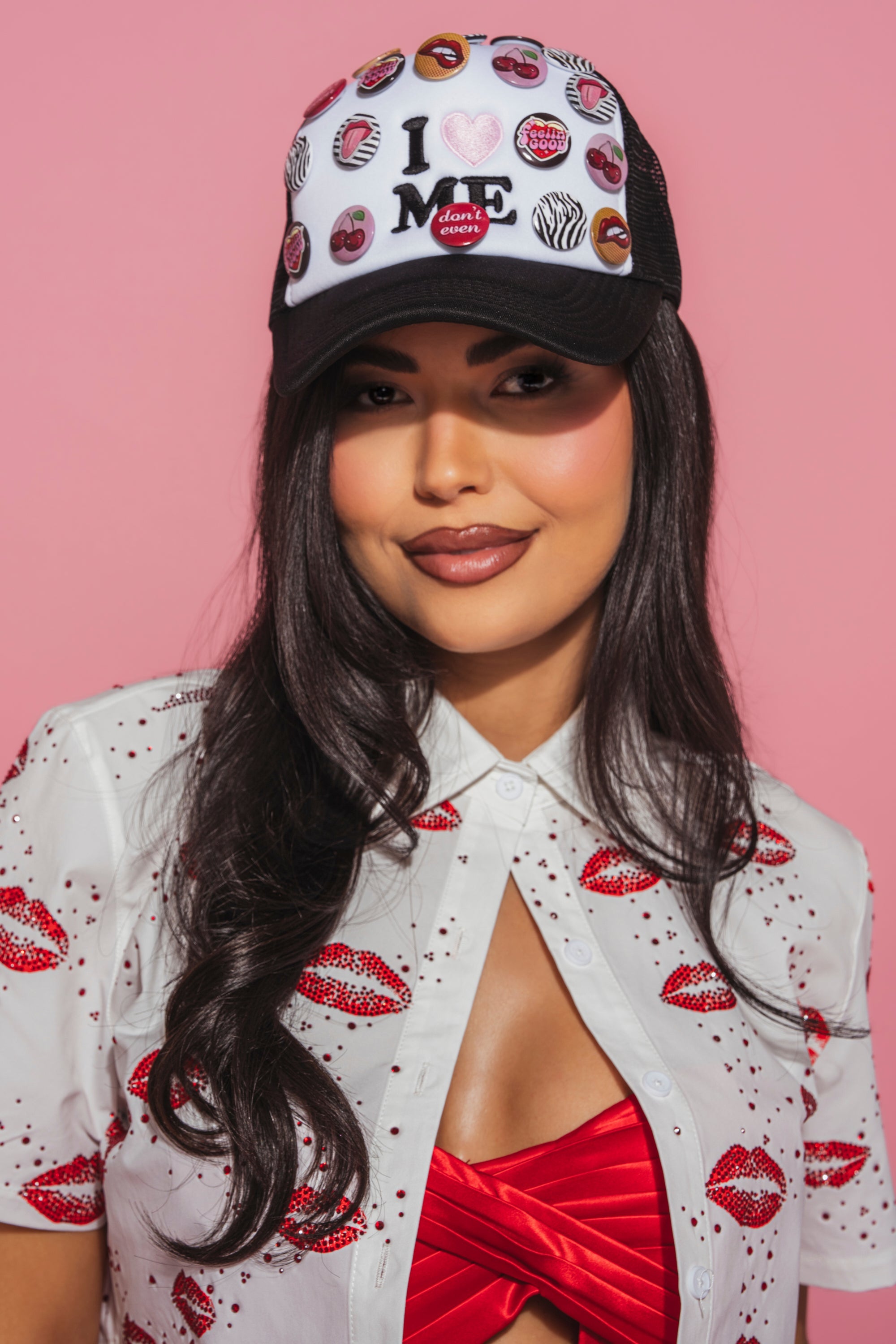 A woman with long dark hair wears a white shirt with red lips prints, a red top underneath, and the LOVE ME BUTTON HAT—a black-and-white trucker hat with candy and heart designs—on a pink background. 1