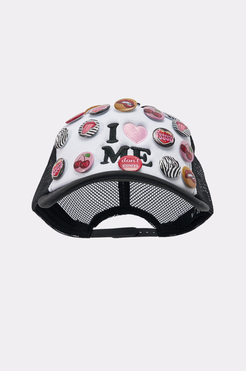 The LOVE ME BUTTON HAT is a black and white mesh-back trucker hat with colorful round pins—cherries, lips, zebra stripes. The front is embroidered with "I ♥ ME" and features a red badge reading "don't worry.