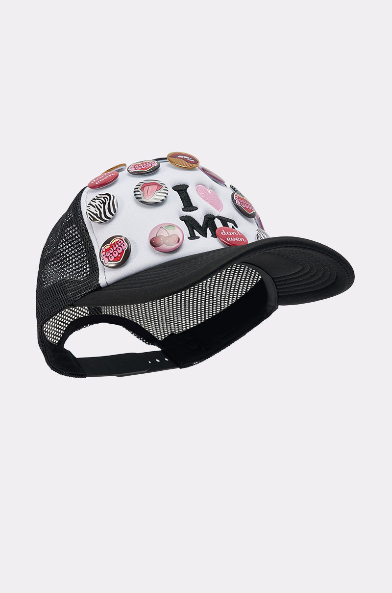 The LOVE ME BUTTON HAT is a black and white mesh-back trucker hat featuring colorful button pins and a bold "I ♥ MCM" design on the front.
