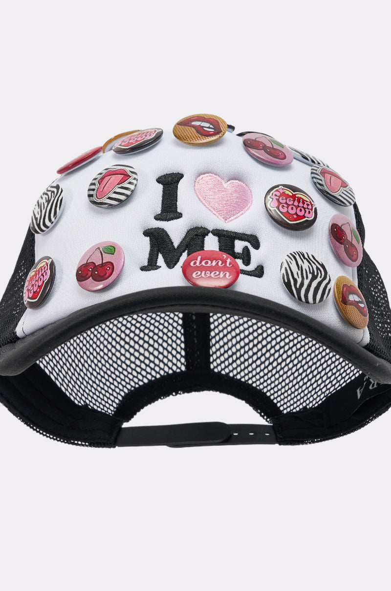The LOVE ME BUTTON HAT is a black and white mesh-back cap with colorful pins—cherries, lips, text—and "I ♥ ME" on the front with a pink heart. A red pin below reads "don't even." Adjustable snapback closure for fit.