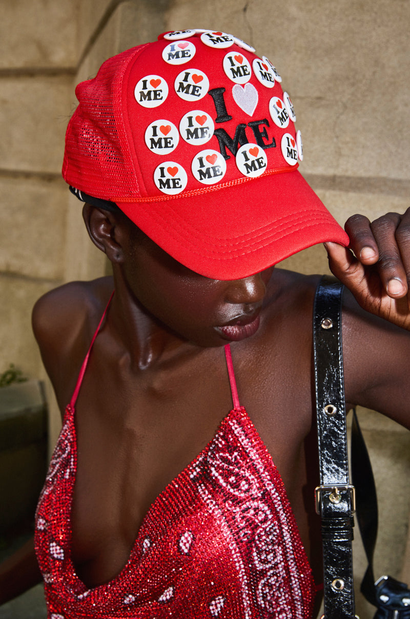 Wearing a red YOU DO TOO PIN HAT and a matching sequined dress, a person looks down while holding the hat’s brim. The background is blurred. 0