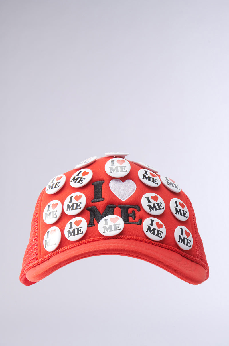 The YOU DO TOO PIN HAT is a red trucker cap with "I ♥ ME" embroidered on the front and adorned with round white pins featuring "I ♥ ME." It has an adjustable snapback for a perfect fit. 1