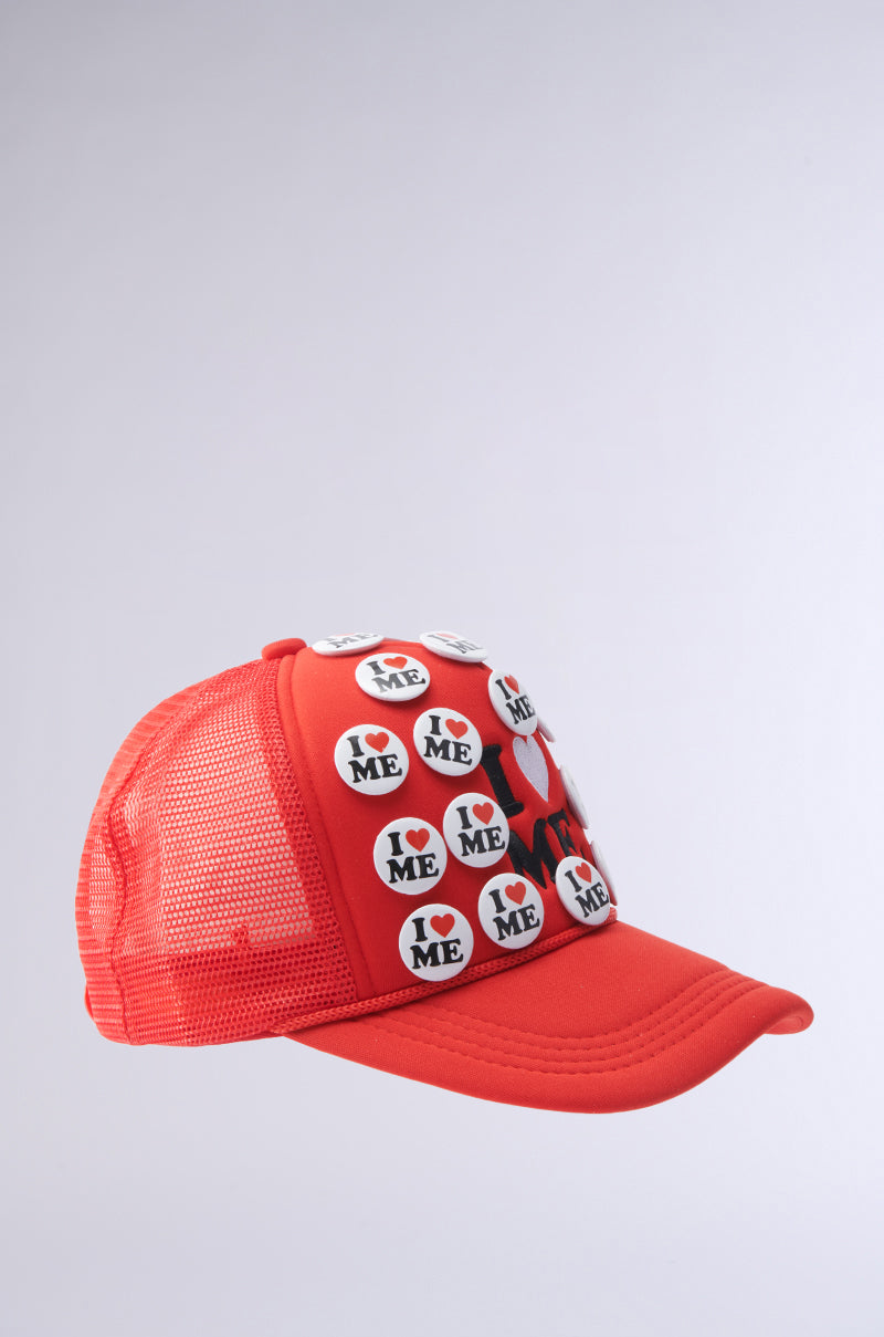 The YOU DO TOO PIN HAT is a red mesh trucker hat with an adjustable snapback, featuring several round pins that say "I ♥ ME" in black text and a red heart, shown on a plain light background. 2