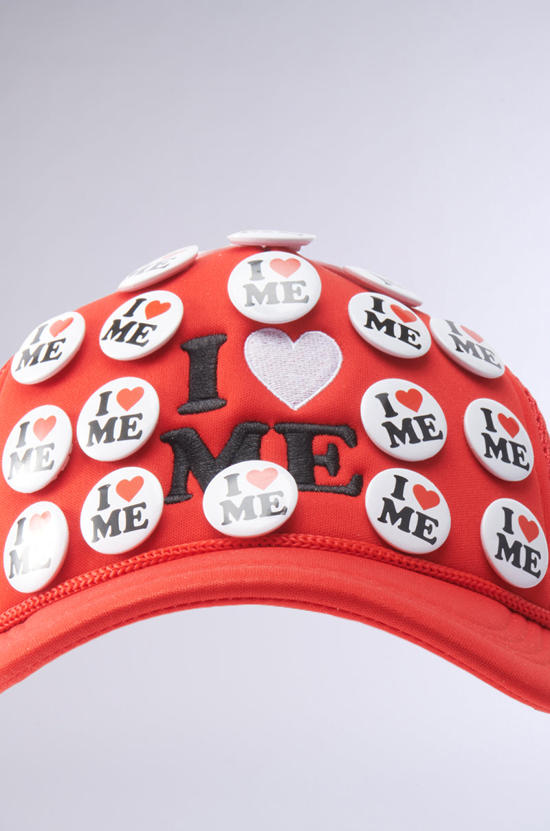 The YOU DO TOO PIN HAT is a red trucker cap with an adjustable snapback, decorated with round "I ♥ ME" buttons and black-and-white "I ♥ ME" embroidery on the front. 3