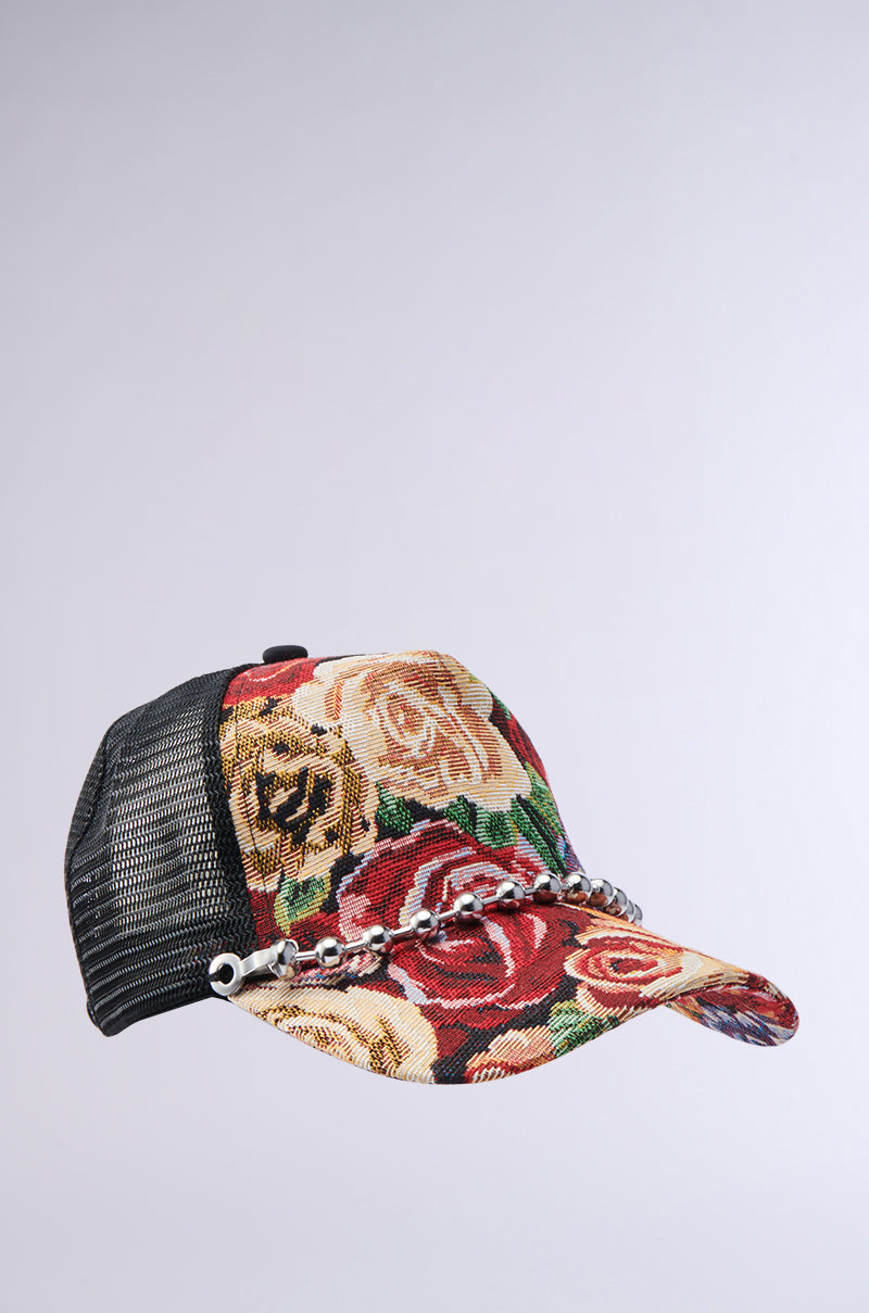 The ROSES ARE RED TAPESTRY HAT is a trucker hat with a mesh back, floral tapestry front and brim featuring red, yellow, and beige roses, and a metal chain detail across the front.