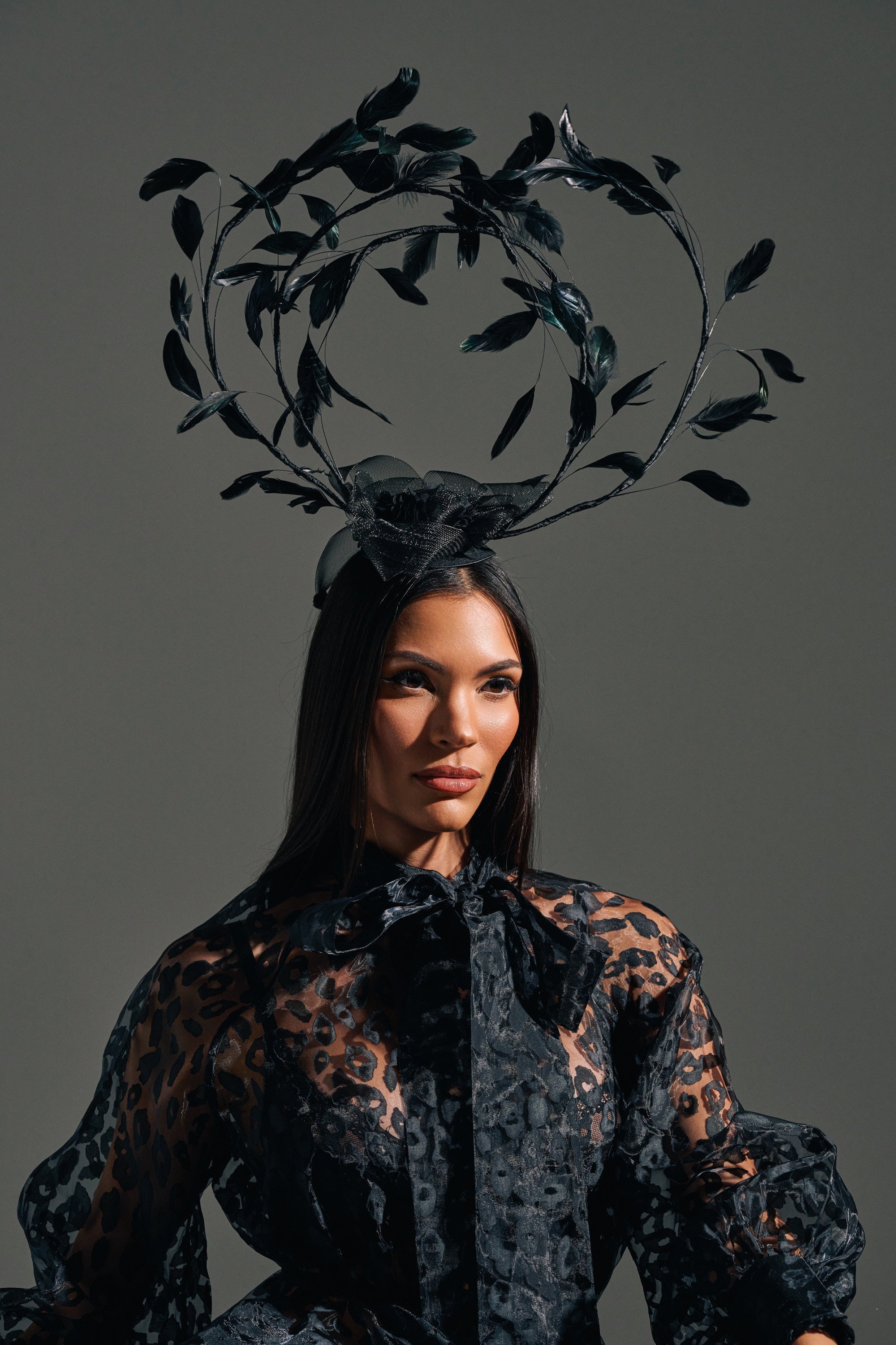 A woman stands against a gray background, wearing the IN YOUR DREAMS CLIP ON HAT with dramatic feathers, swirling branches, and leaves. She has long dark hair and pairs the hat with a sheer patterned black top featuring a bow at the neck. 0