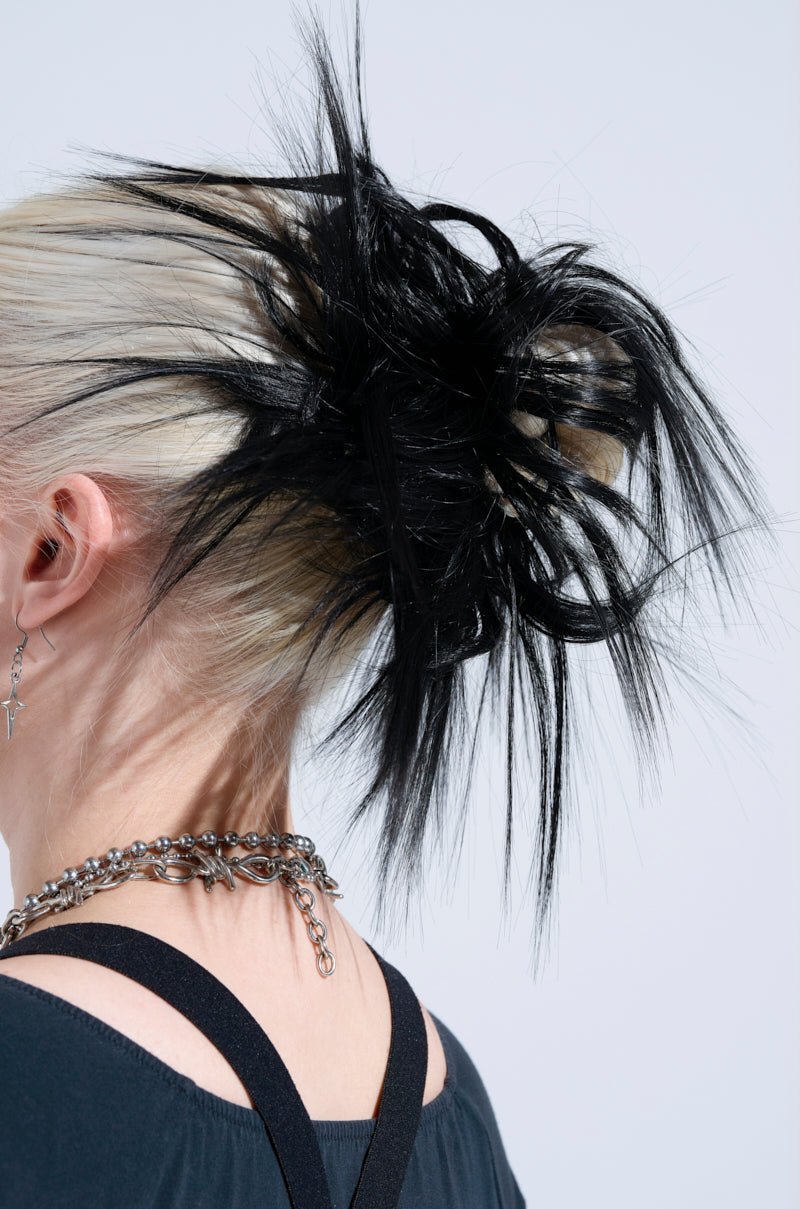 The person rocks a punk-glam style with platinum blonde hair in a voluminous updo, highlighted by the SPIKED OUT HAIR PIECE. A black top complements a silver chain necklace and cross earring, crafting an edgy look against the light backdrop.