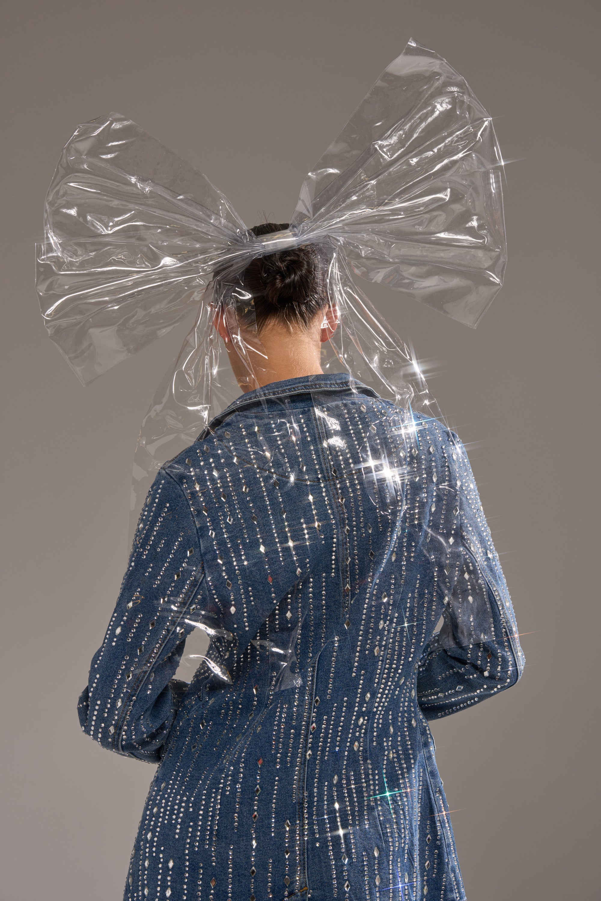A person is seen from behind wearing a sparkling blue jacket and the CLEARLY A MOMENT OVERSIZED BOW, a large translucent PVC bow secured with an alligator clip, set against a neutral gray background.