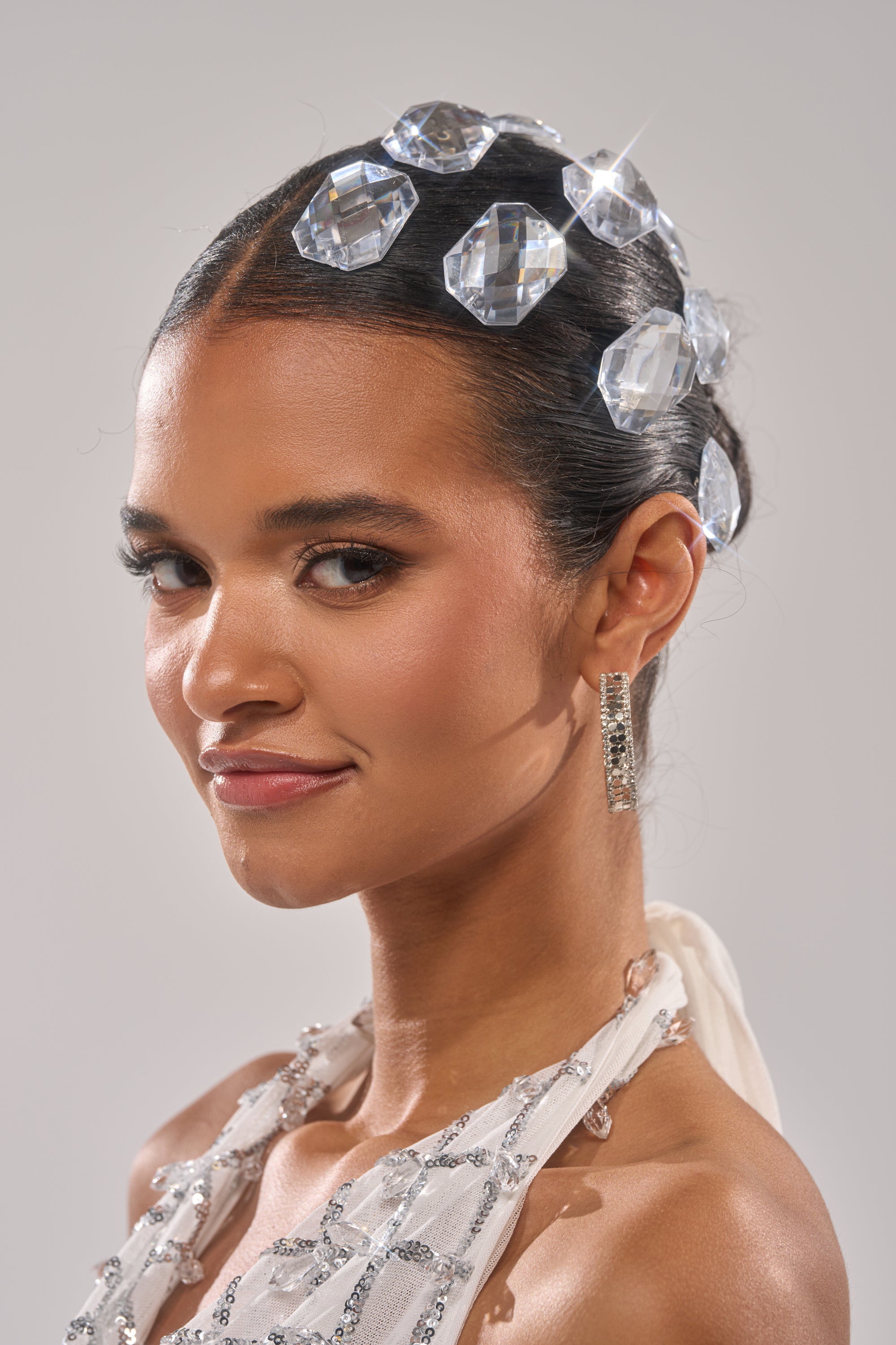 A woman with sleek dark hair wears the DIAMONDS DANCING HAIR CLIP SET, featuring sparkling barrette clips and gem-like crystals. She pairs natural makeup, dangling earrings, and a white embellished outfit against a plain background.