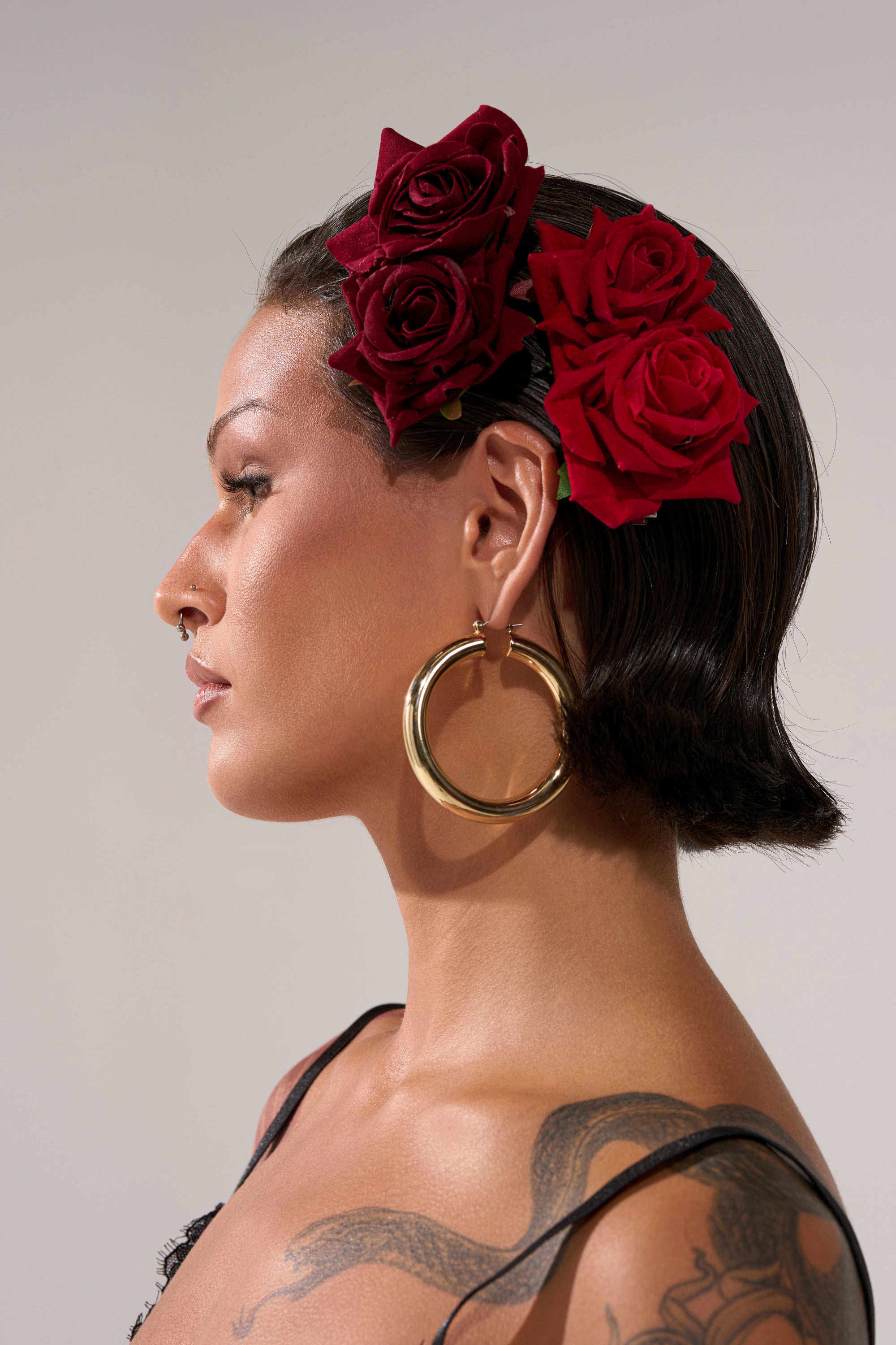 A person with slicked-back dark hair, large gold hoop earrings, and visible shoulder tattoos wears a sleek black top and tucks ROSES ARE RED HAIR CLIPS behind their ear as a bold statement.