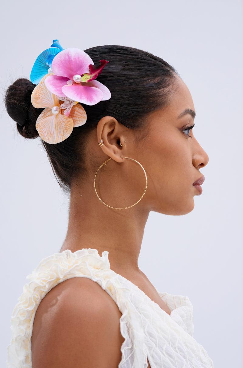 Woman in profile with hair in a bun adorned with the INTO THE GARDEN HAIR CLIP SET—colorful tropical flowers, pearls, and gold clips—plus large gold hoop earrings and a textured white sleeveless top, set against a plain light background.