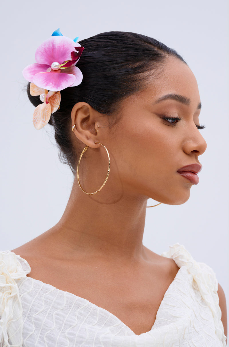A woman with smooth, dark hair in a bun, adorned with an INTO THE GARDEN HAIR CLIP SET in pink and purple. She wears gold hoop earrings and a textured white top, gazing serenely to the side against a plain background. 1