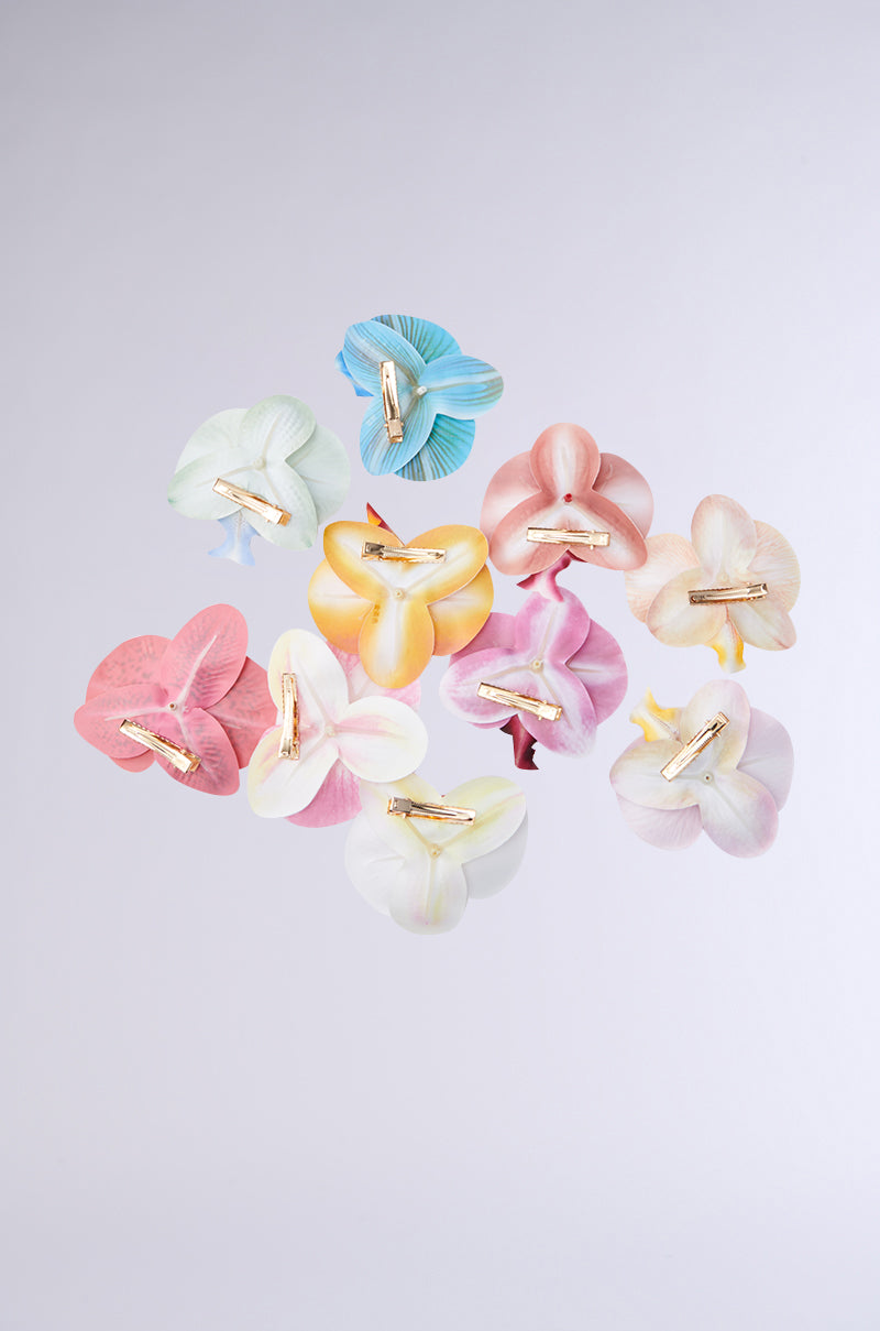 The INTO THE GARDEN HAIR CLIP SET features ten pastel tropical flower clips with metallic gold hardware, arranged in a circle on a light gray background for an elegant touch. 3