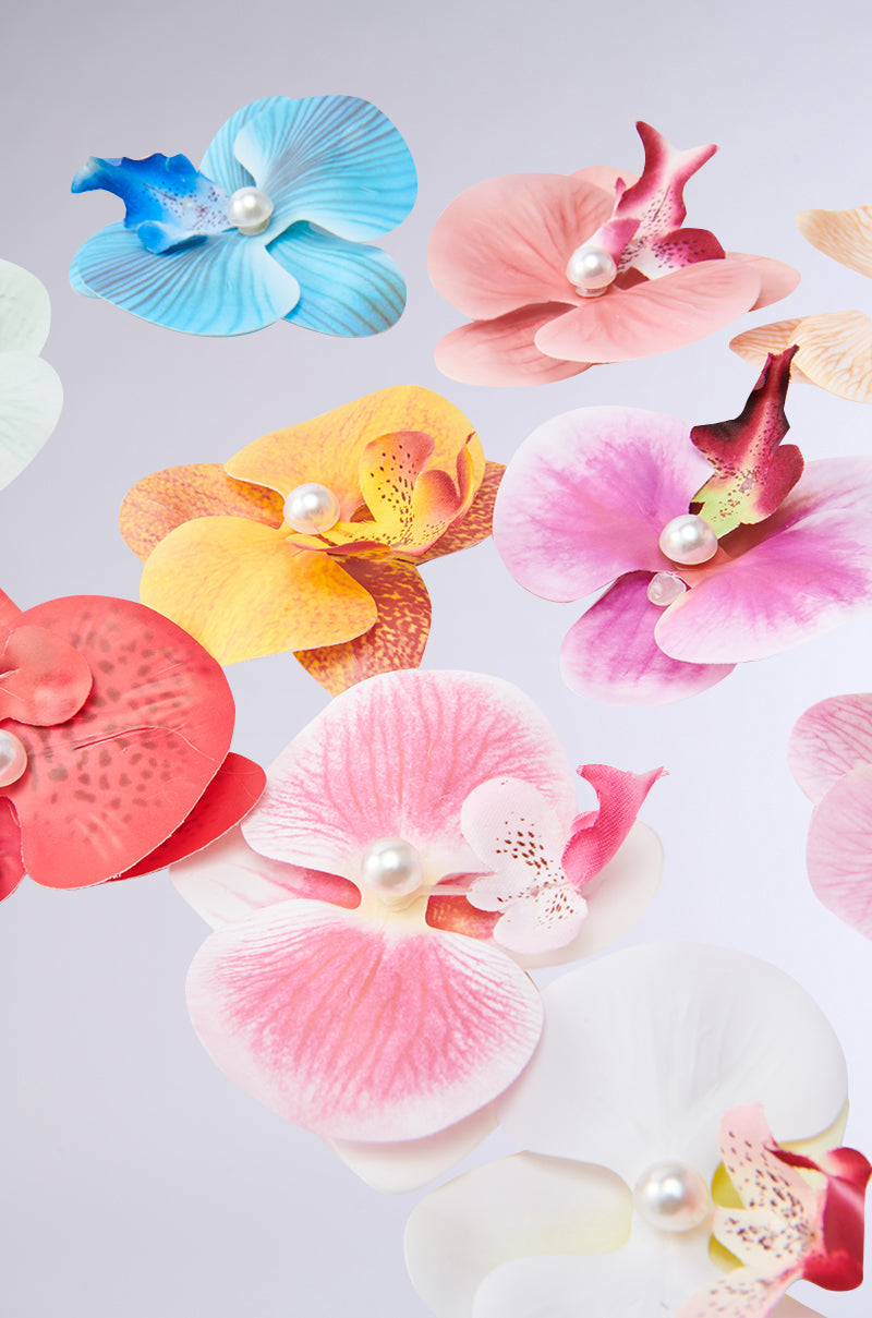 Colorful artificial orchids with pearl centers are scattered on a light gray background, resembling the vibrant INTO THE GARDEN HAIR CLIP SET in a delicate and eye-catching display.