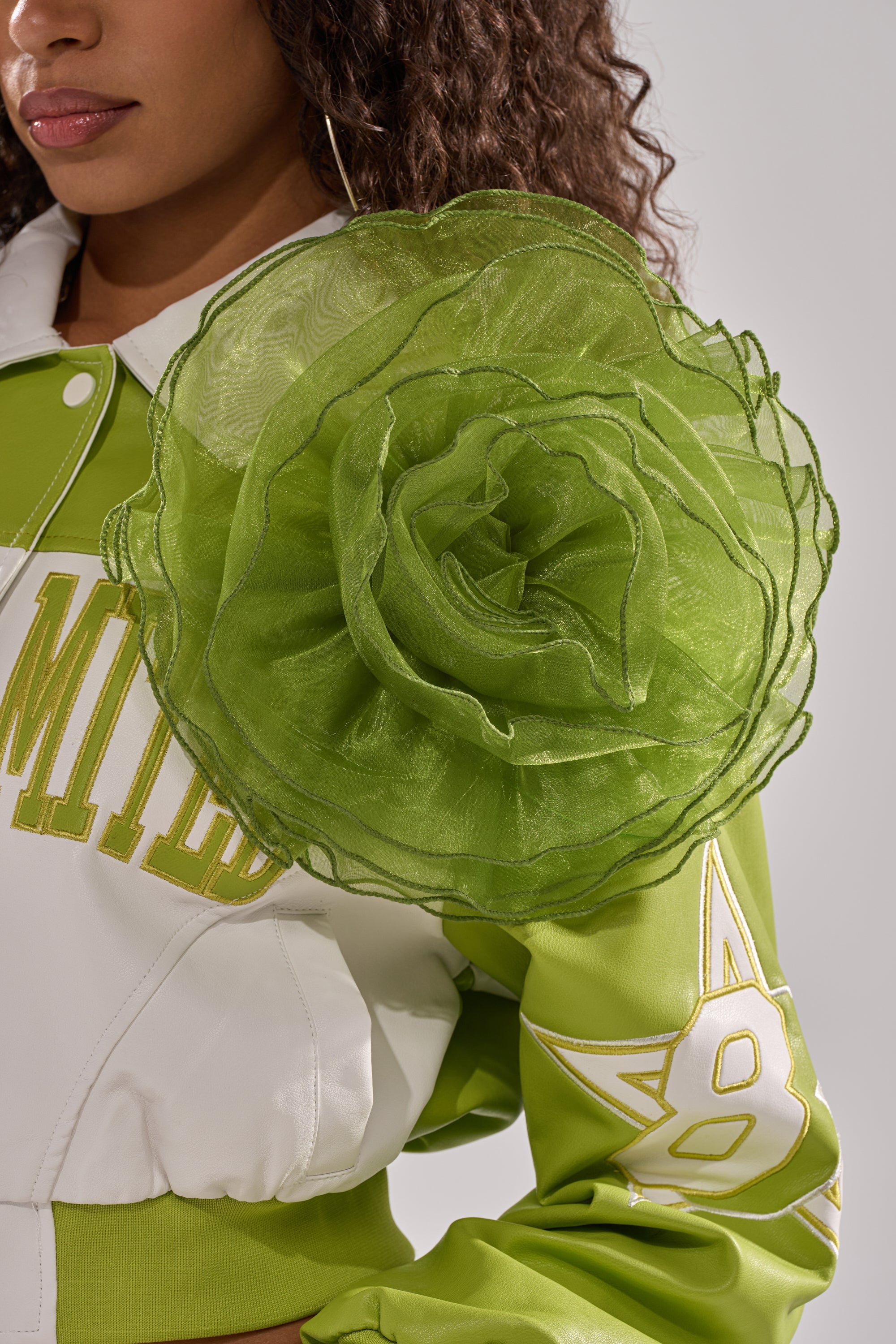 A person wears a green and white jacket with the NO ENVY BROOCH, an oversized sheer organza floral accessory, on the shoulder. Only the lower half of their face and upper body are visible. 0