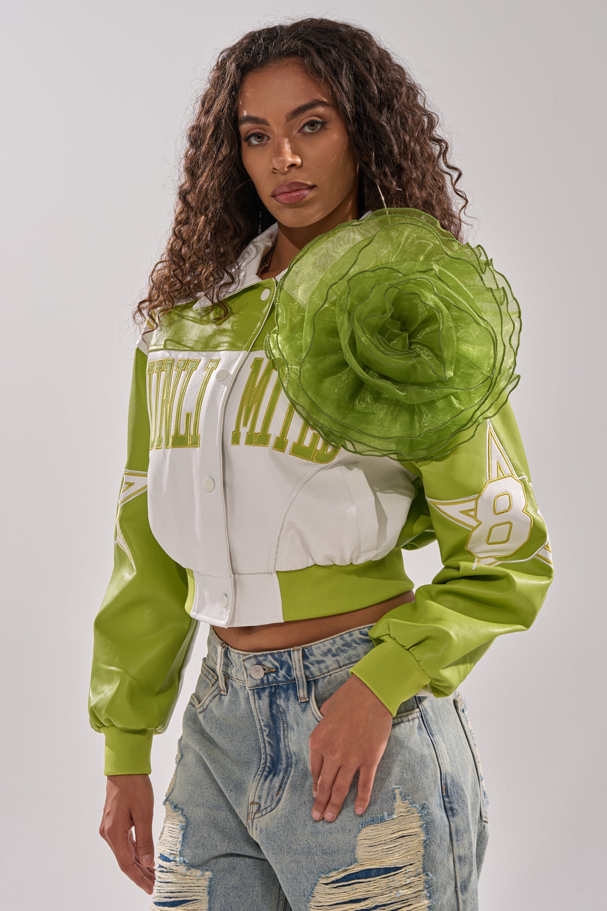 A woman with long curly hair wears a green and white varsity jacket, distressed denim jeans, and the NO ENVY BROOCH on her shoulder, standing against a plain background. 1