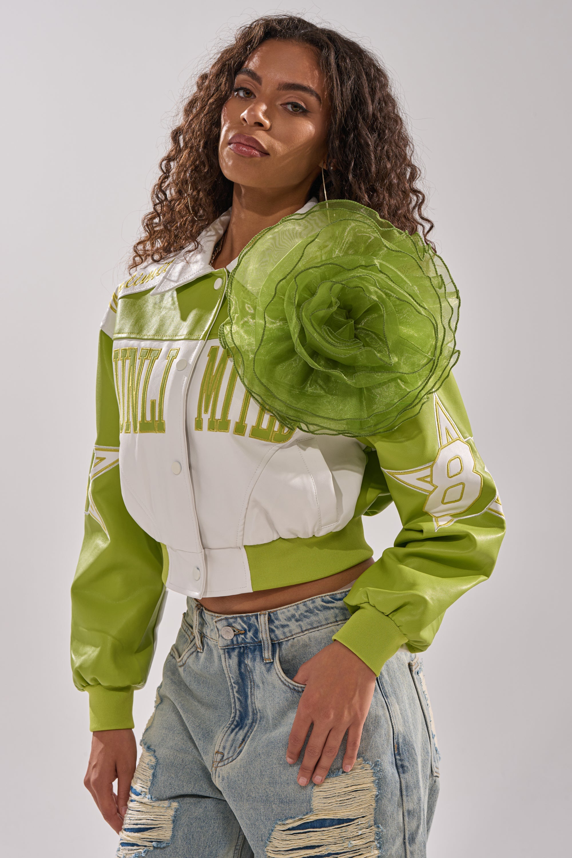 A woman with long curly hair wears a white and green varsity-style jacket adorned with the NO ENVY BROOCH on her shoulder, paired with light blue ripped jeans, standing against a plain background. 2