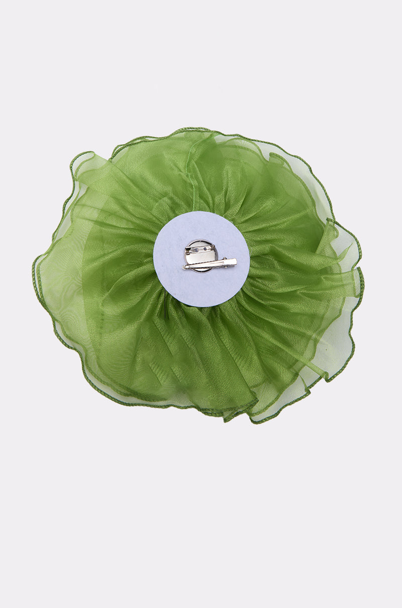 The NO ENVY BROOCH features a green sheer fabric flower with ruffled edges, set on a round white base with a silver metal clasp. This AKIRA Label piece doubles as an oversized hair clip or brooch, shown on a plain white background. 4