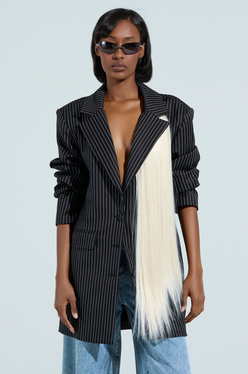 A person in a pinstripe blazer, featuring safety pin closures and the NOT FOR THE WEAK SYNTHETIC HAIR BROOCH, stands against a light gray background. They wear sunglasses, have partially visible chest under the buttoned blazer, and are styled with blue jeans.