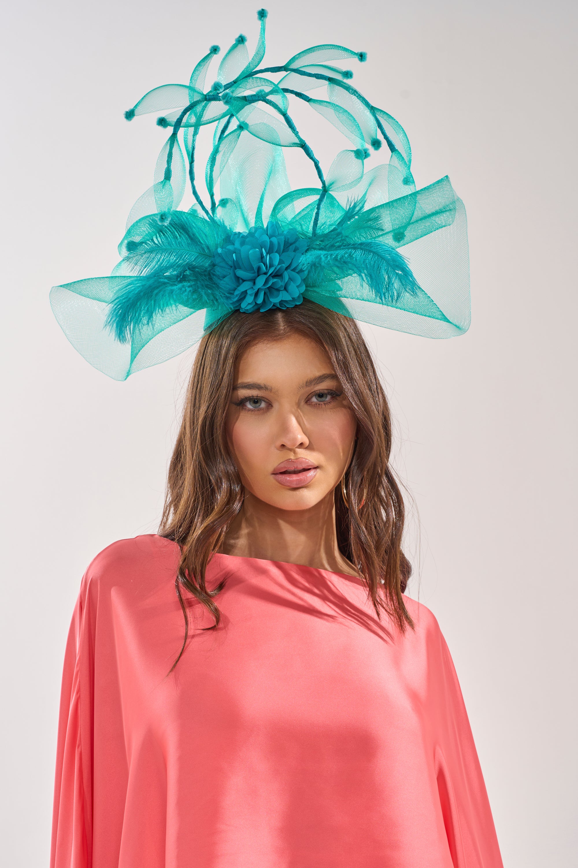 A woman poses against a plain light background wearing a bright coral pink dress and the INTO THE GARDEN BLOOMING HAT, an elaborate turquoise fascinator with netting, feathers, and decorative loops. 0
