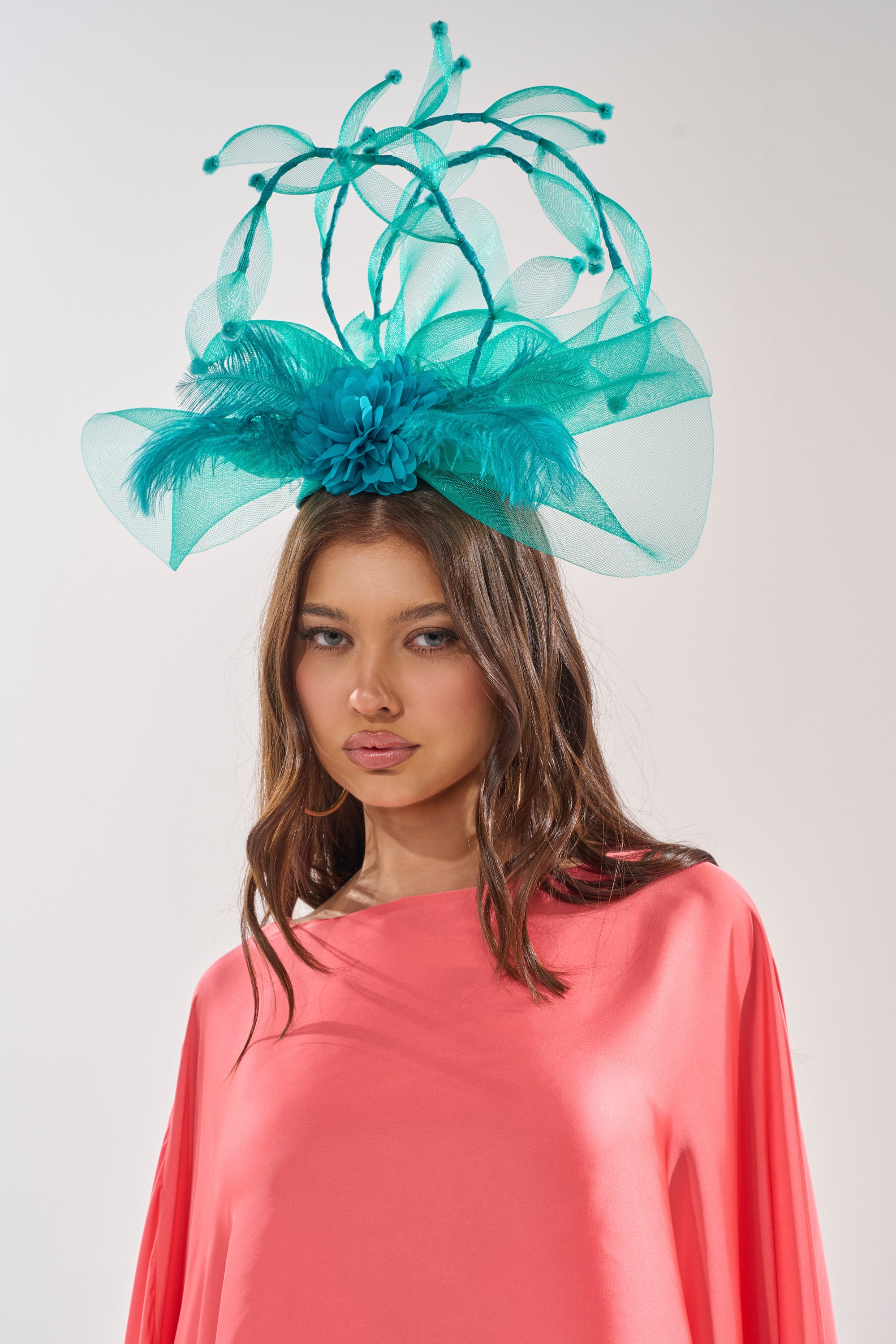 A woman in a coral pink outfit showcases the INTO THE GARDEN BLOOMING HAT, an elaborate turquoise feathered fascinator with mesh and decorative spikes—ideal as a standout race day headpiece or wedding accessory. 1
