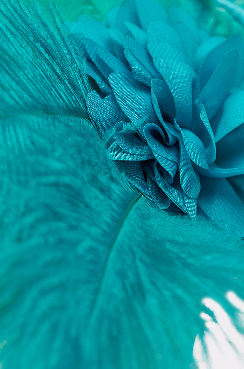 Close-up of the INTO THE GARDEN BLOOMING HAT shows its vibrant turquoise feather fascinator and matching fabric flower, highlighting their detailed textures—an elegant accessory perfect for weddings or any formal occasion. 4