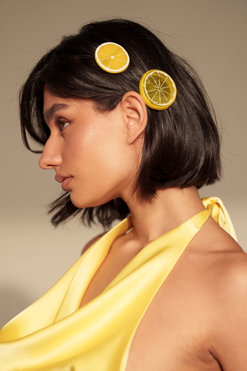 A woman with short dark hair wears the LIFE GIVES YOU LEMONS HAIR CLIP SET—barrette-style clips shaped like lemon and lime slices—with a yellow satin halter top, photographed in profile against a neutral background. 0