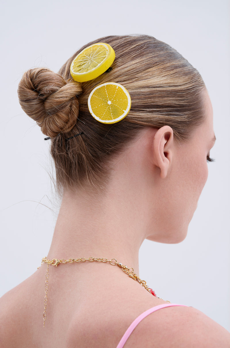 A woman with blonde hair in a neat bun, styled with the LIFE GIVES YOU LEMONS HAIR CLIP SET—lemon and orange-shaped barrettes—wears a gold chain necklace and pink strap top, seen from the back against a plain light background. 1
