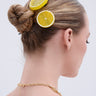 A woman with blonde hair in a neat bun, styled with the LIFE GIVES YOU LEMONS HAIR CLIP SET—lemon and orange-shaped barrettes—wears a gold chain necklace and pink strap top, seen from the back against a plain light background.