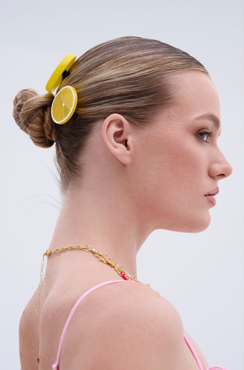 A woman with blonde hair in a bun wears the LIFE GIVES YOU LEMONS HAIR CLIP SET as a playful accessory, paired with a gold chain necklace and a pink top, standing against a light background. 2