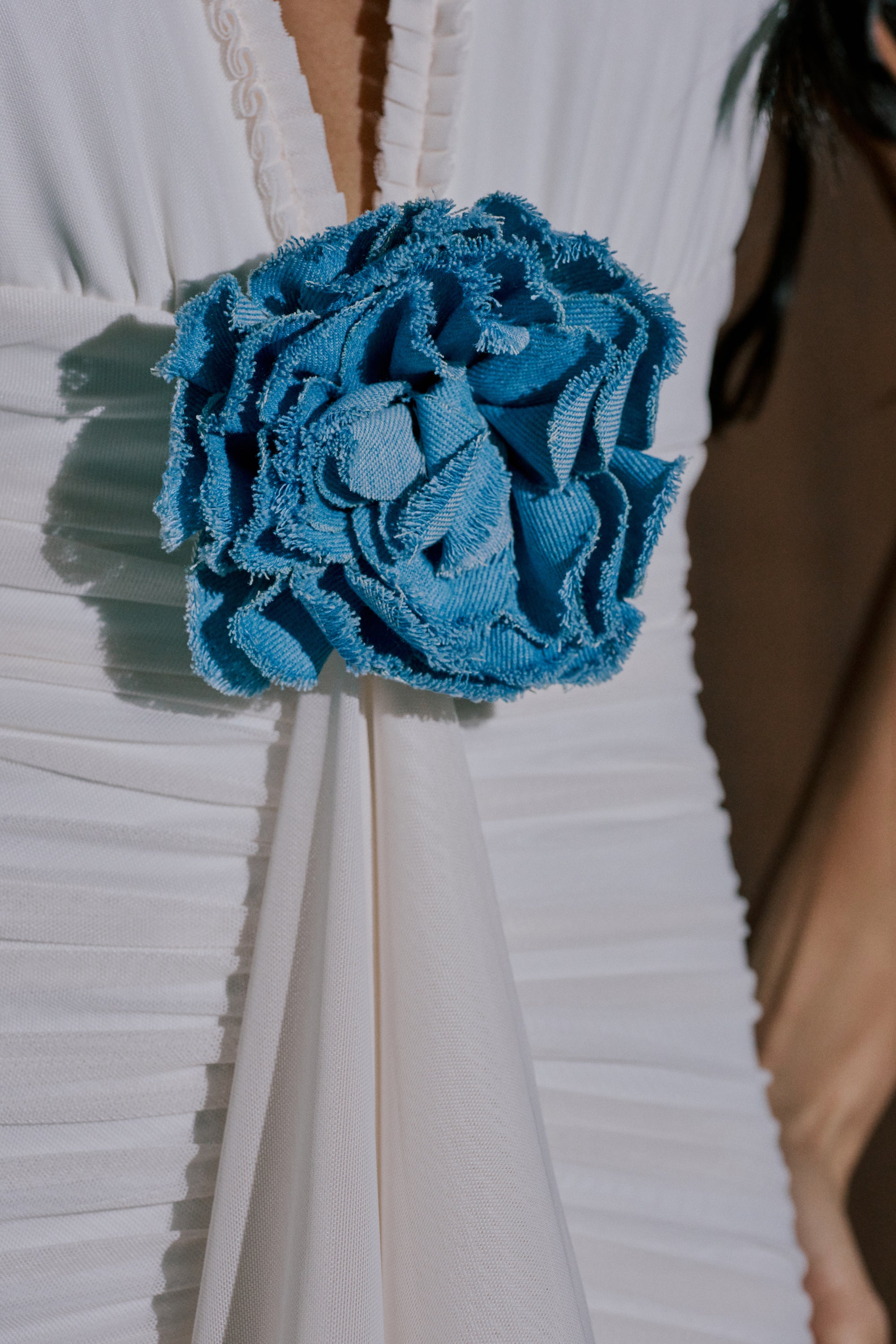 A close-up of a white pleated dress adorned with the DENIM ERA BROOCH—a large, textured blue denim flower at the waist—adding a stylish, modern accent. 3