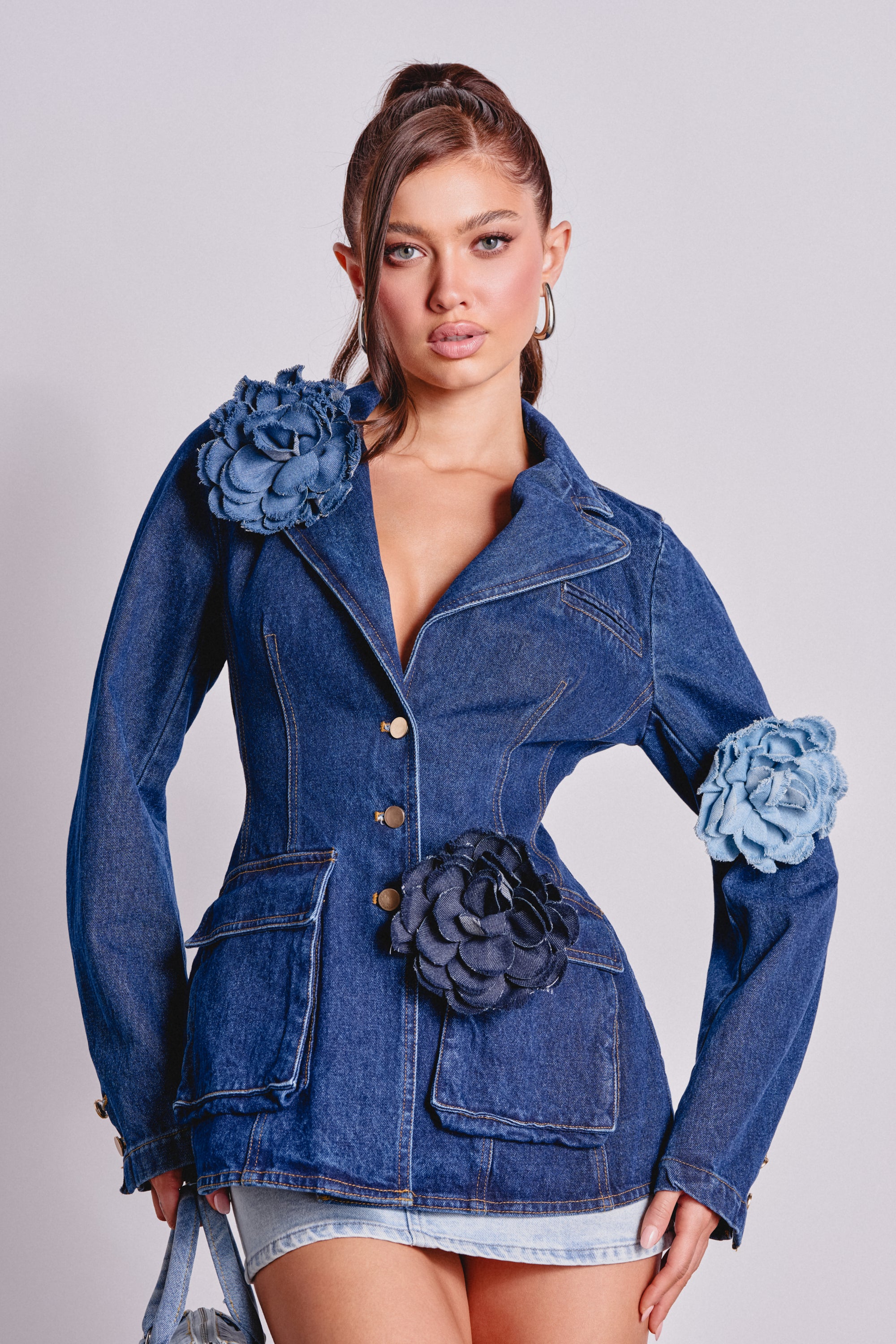 A woman showcases the DENIM ERA BROOCH—a striking denim blazer with oversized fabric flower appliqués on the shoulder, waist, and sleeve—styled with a high ponytail and denim shorts. 0