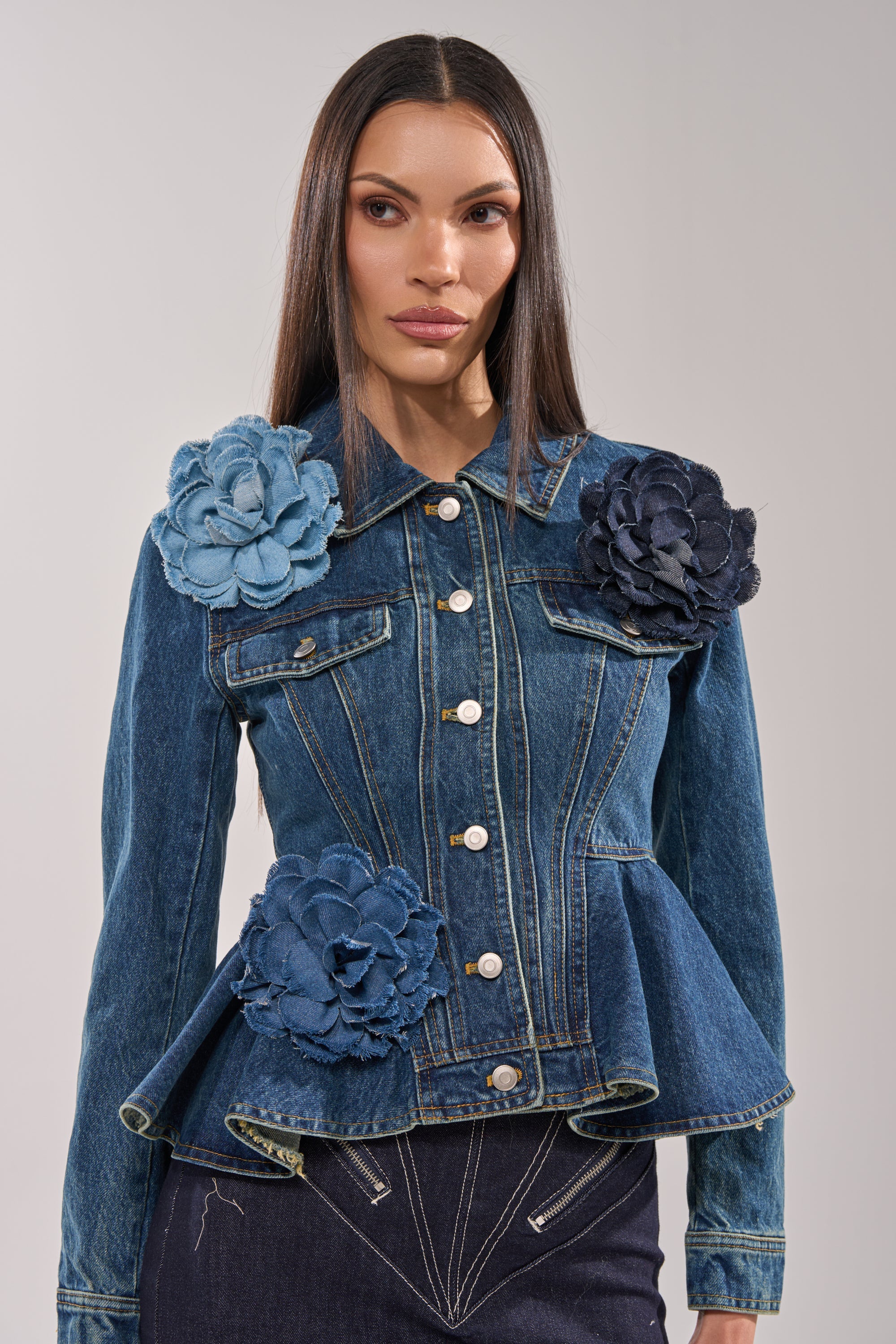 A woman with straight, dark hair and a neutral expression wears a fitted denim jacket with silver buttons and a flared hem, accessorized with the DENIM ERA BROOCH as an oversized floral pin on her shoulder and hip. 5