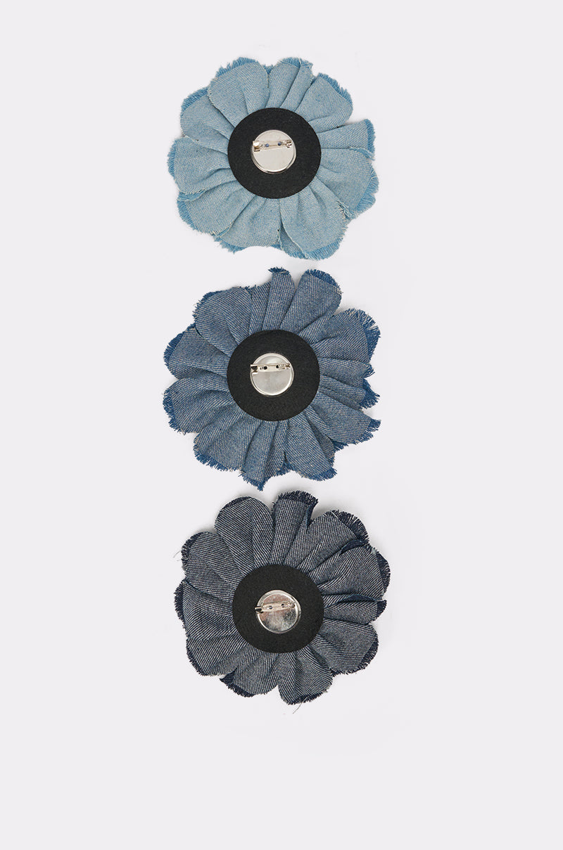 The DENIM ERA BROOCH set includes three vertically arranged denim flower brooches in light, medium, and dark blue with frayed edges, black centers, and pin backs, all displayed on a crisp white background. 7