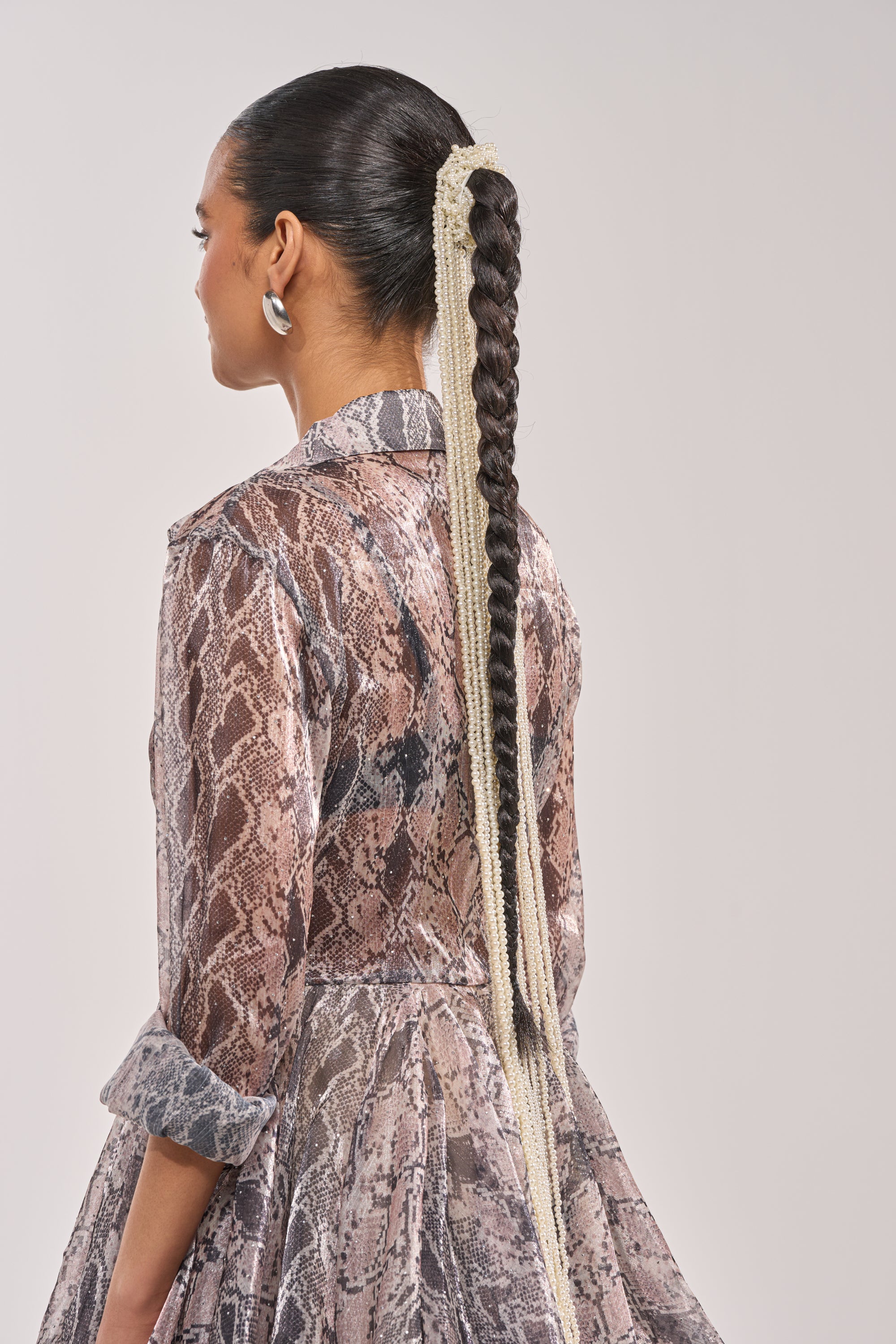 A person with long, dark hair in a sleek braid, secured by the PURE VIBES PEARL HAIR ACCESSORY, wears a sheer snakeskin-print dress with rolled-up sleeves and faces away from the camera against a plain background. 0