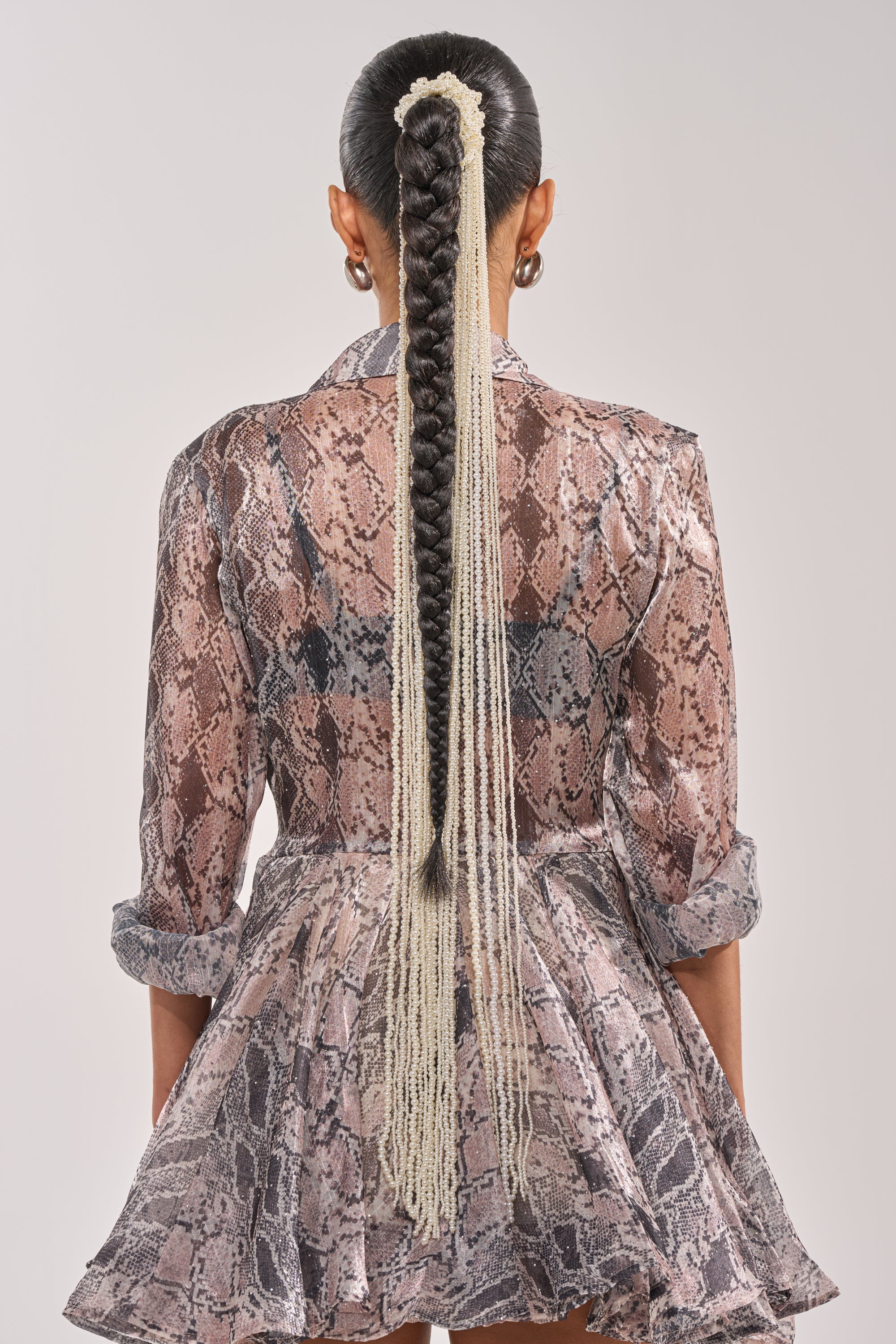 A person with long, dark braids styled with the PURE VIBES PEARL HAIR ACCESSORY stands facing away, wearing a sheer, patterned dress featuring a flared skirt and rolled-up sleeves. 1