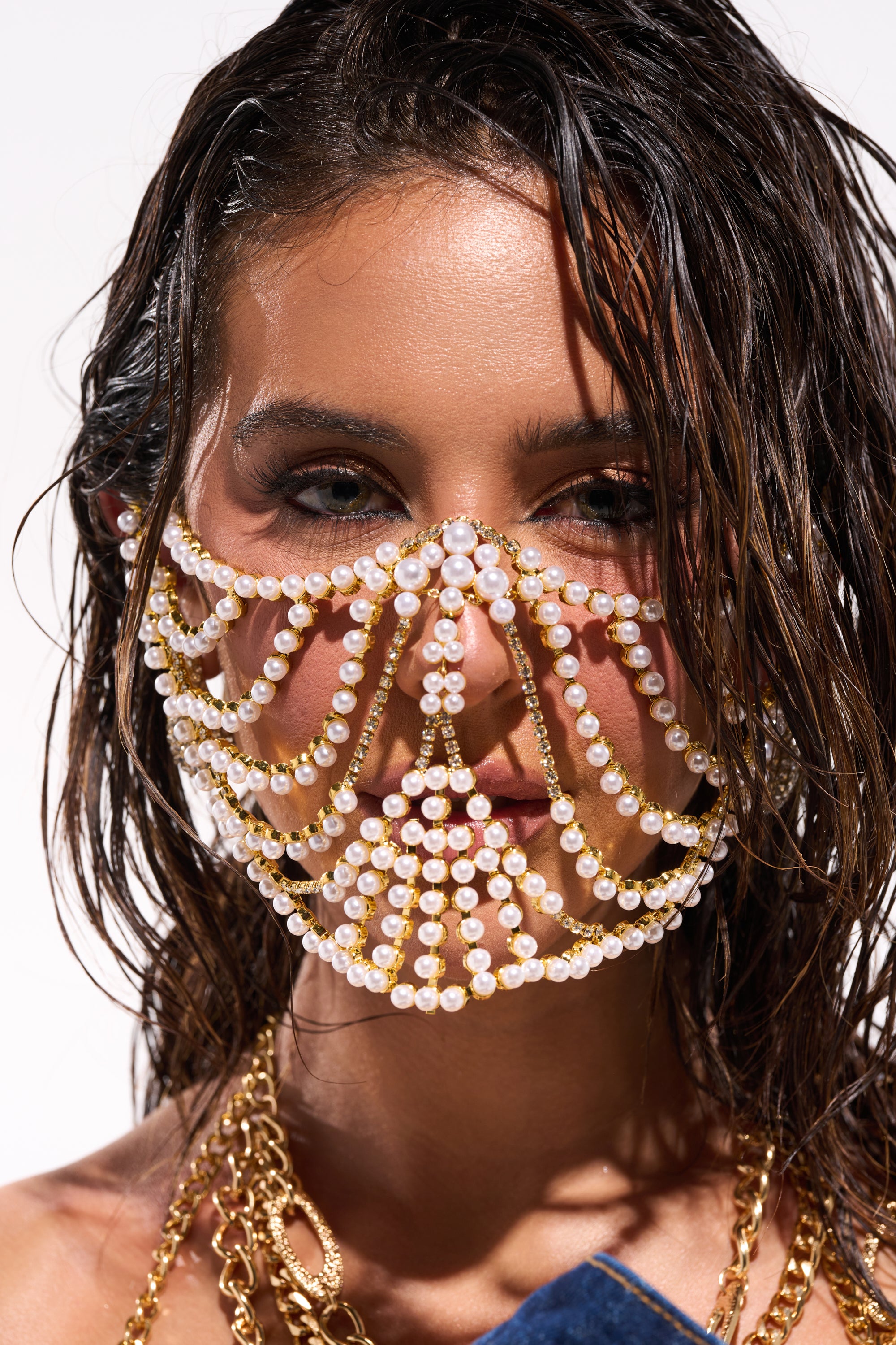 A woman with wet hair wears the DONT TALK PEARL MOUTHPIECE, a decorative festival face jewelry chain featuring pearls and gold links draped across her face. She pairs it with gold necklaces and looks directly at the camera. 0