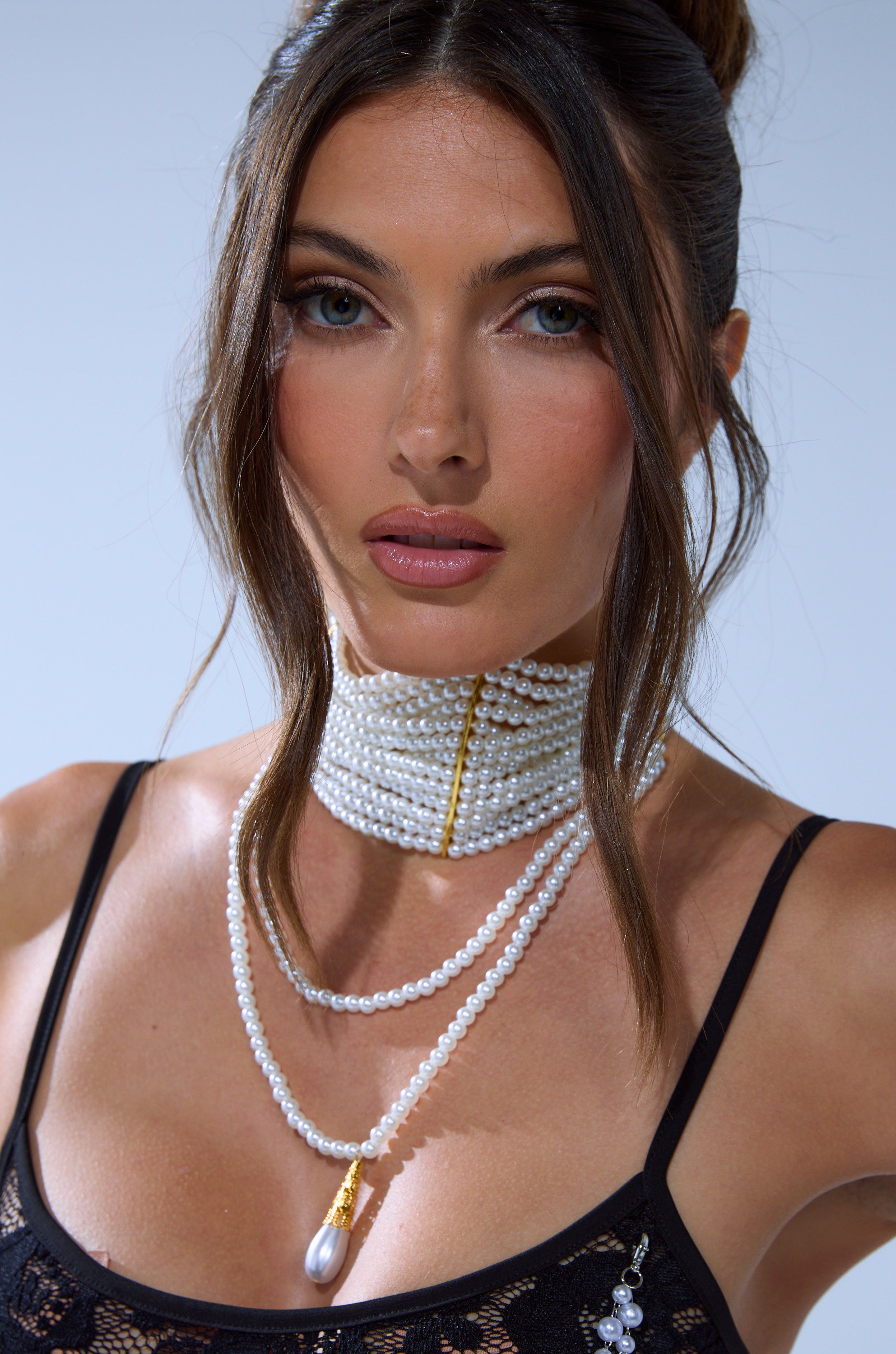 A woman with long brown hair wears a black lace top and the VERSAILLES NECKLACE, a layered pearl choker that makes a perfect statement accessory. She has natural makeup and a neutral expression.