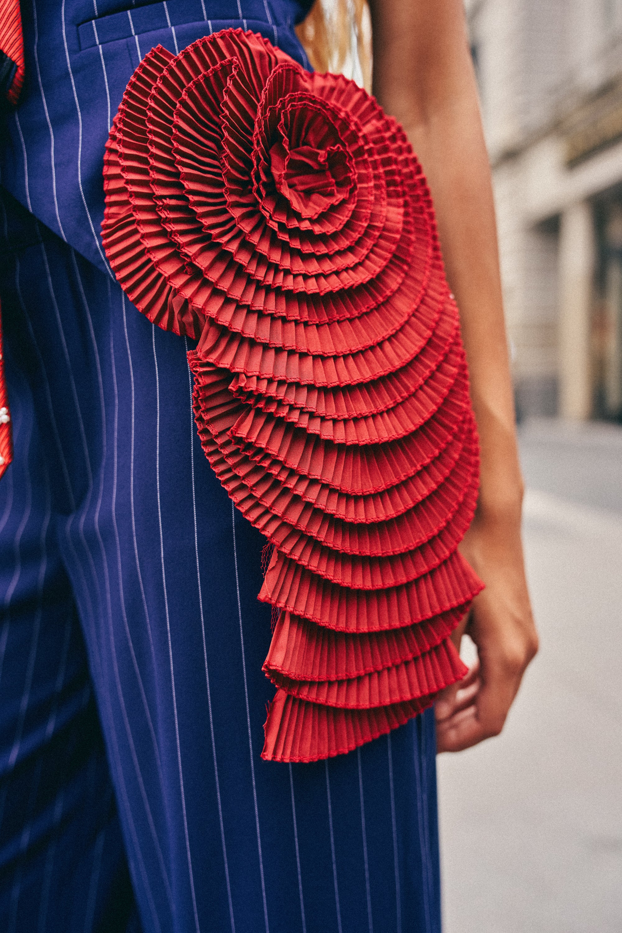 A person wears blue pinstripe pants adorned with the IN BLOOM BROOCH—a large, pleated satin accessory in vivid red, sculpted to resemble a flower or seashell—pinned at the hip. The background reveals an urban street scene.