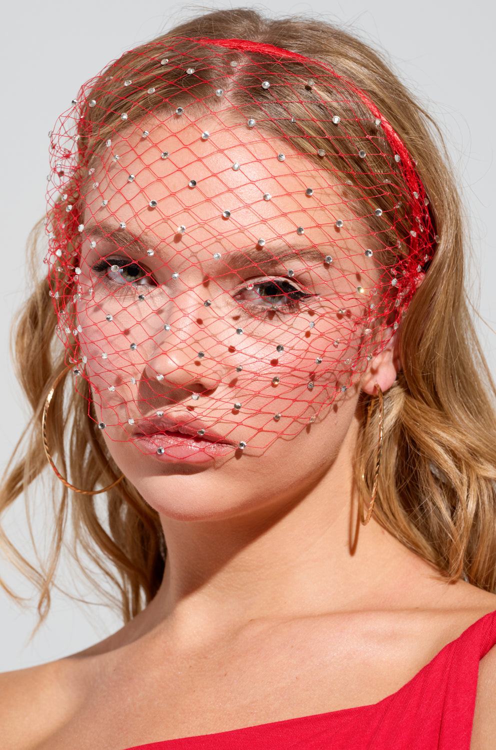 A woman with long blonde hair wears the I SEE FIRE RHINESTONE HEADBAND, a red fishnet mesh veil adorned with small gems. She sports large hoop earrings and a red off-the-shoulder top against a light gray background. 2