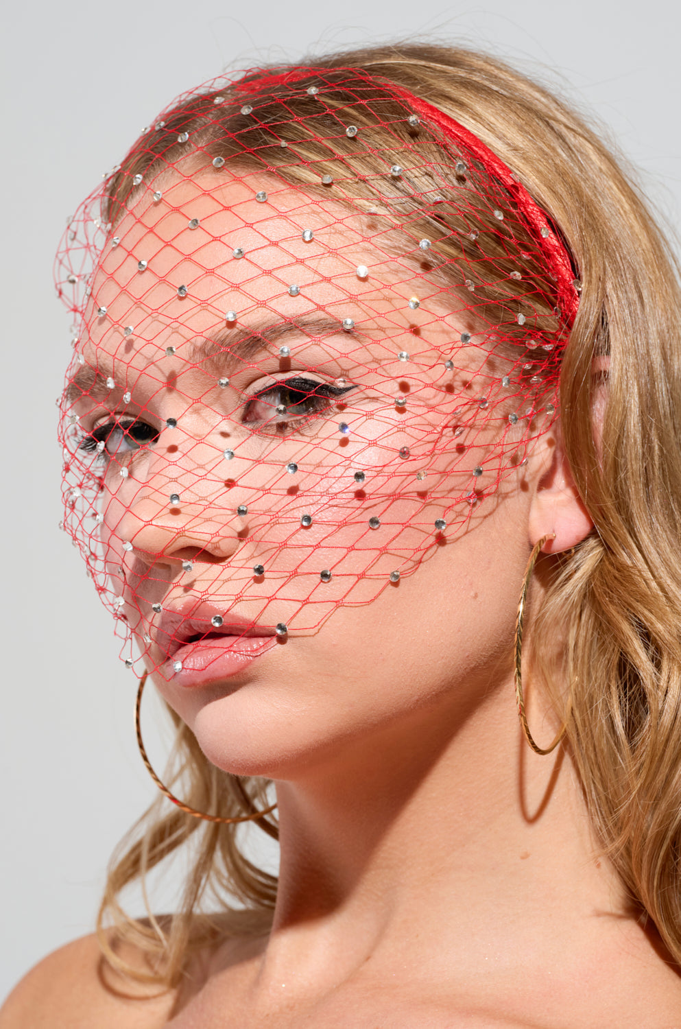 A person with long hair wears a striking red fishnet mesh veil adorned with tiny jewels. Paired with hoop earrings and the I See Fire Rhinestone Headband, they gaze directly at the camera, embodying an effortlessly elegant style reminiscent of the AKIRA Label's signature flair. 0